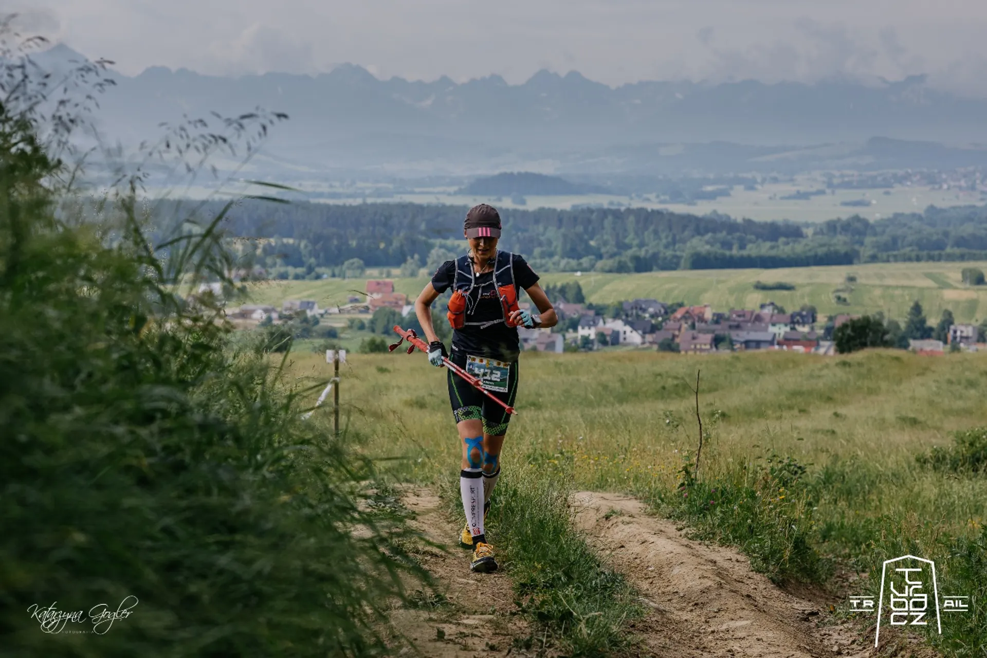The image depicts a trail runner in motion. The runner is wearing a cap, sunglasses, a tank top, shorts, calf compression sleeves, and running shoes equipped for trail running. They are carrying a hydration pack on their back, which often includes storage for water and possibly nutrition or other essentials needed during a long-distance run.

The runner is on a dirt path with grass on either side, indicating a natural outdoor setting. In the background, there is a vast open landscape with a view of a small town or settlement, fields, and a majestic range of mountains in the far distance, which provides a scenic backdrop for the trail run.

The weather appears to be overcast, as suggested by the cloudy sky, which might provide cooler conditions for the runner