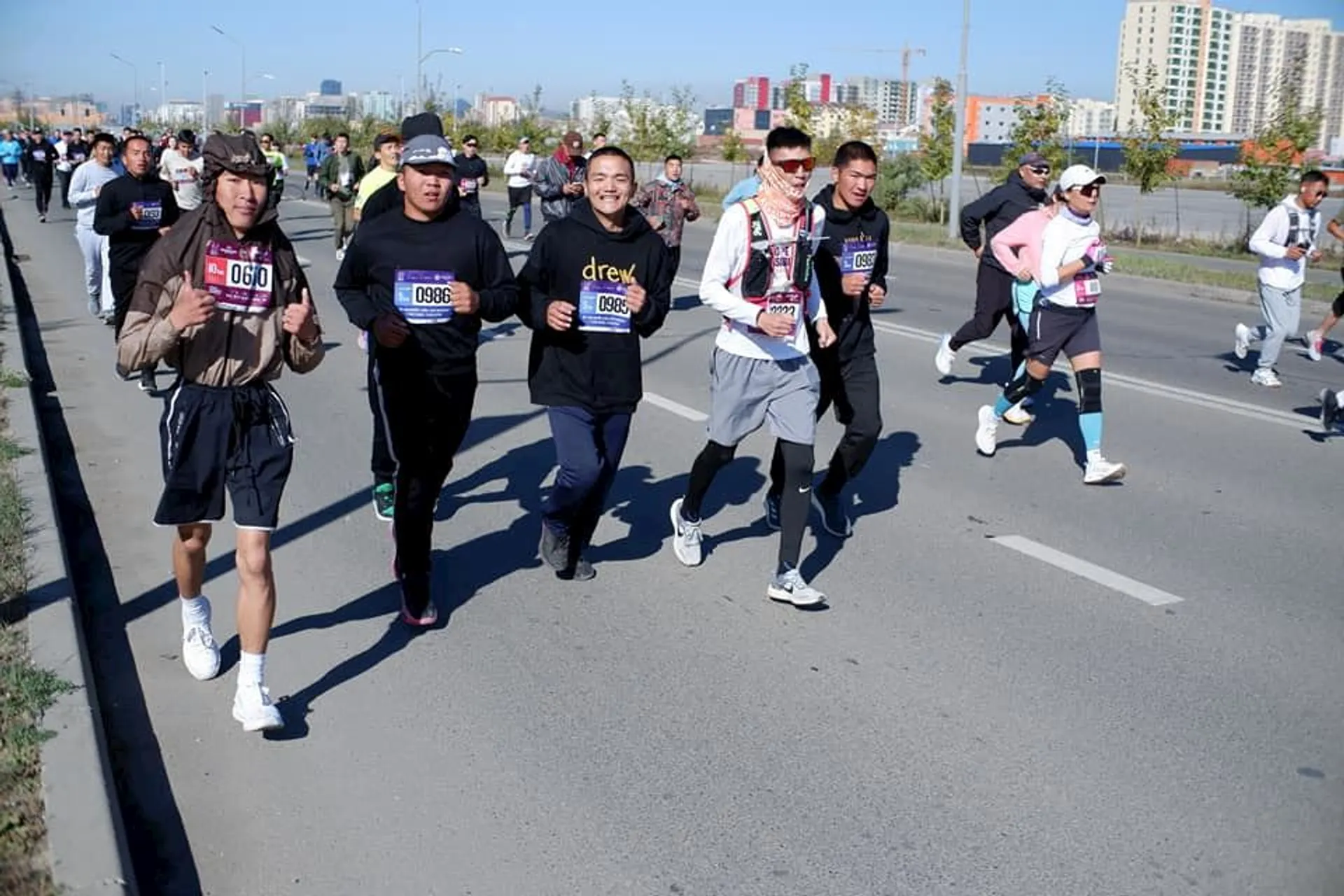 The image shows a group of people running in what appears to be a marathon or organized race. They are on a road, wearing athletic clothes and bib numbers. There are buildings and trees in the background.