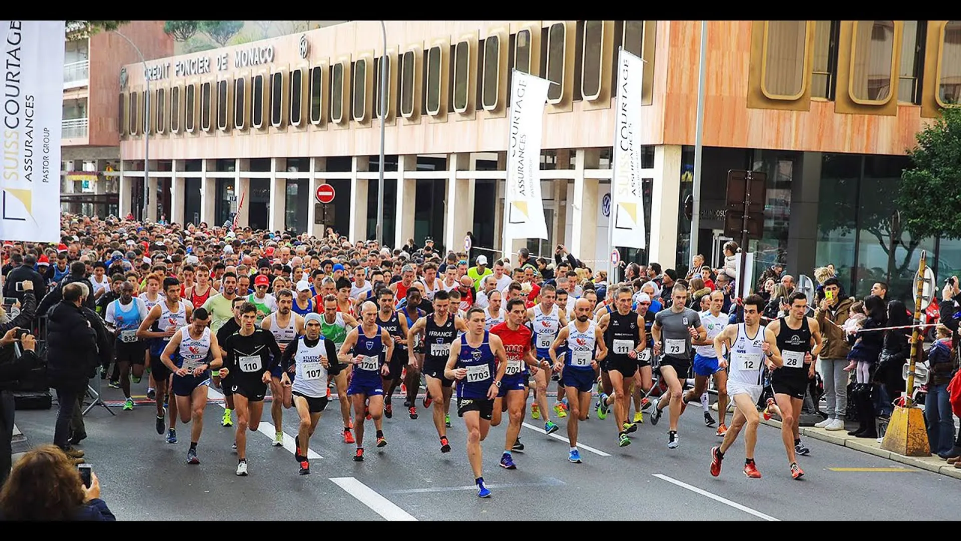 The image shows a large group of runners at the start of a road race.