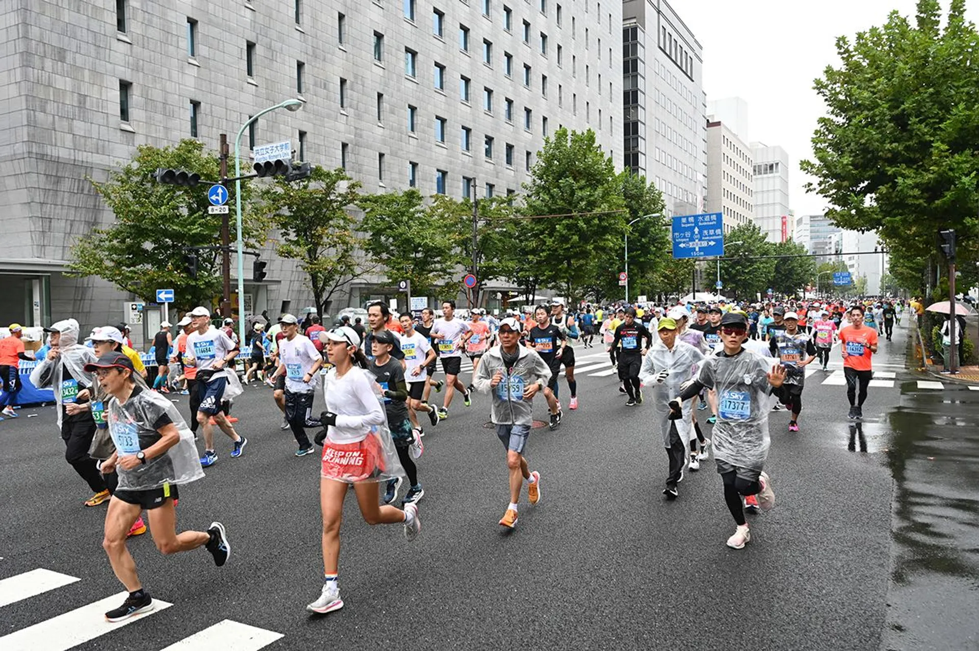 The image shows a group of people participating in a road race or marathon. They are running on a street lined with buildings and trees. Some runners are wearing rain gear, indicating it might be rainy or wet. There are signs and markings for the event, and many participants are dressed in athletic gear like shirts, shorts, and running shoes. The atmosphere appears lively and energetic.
