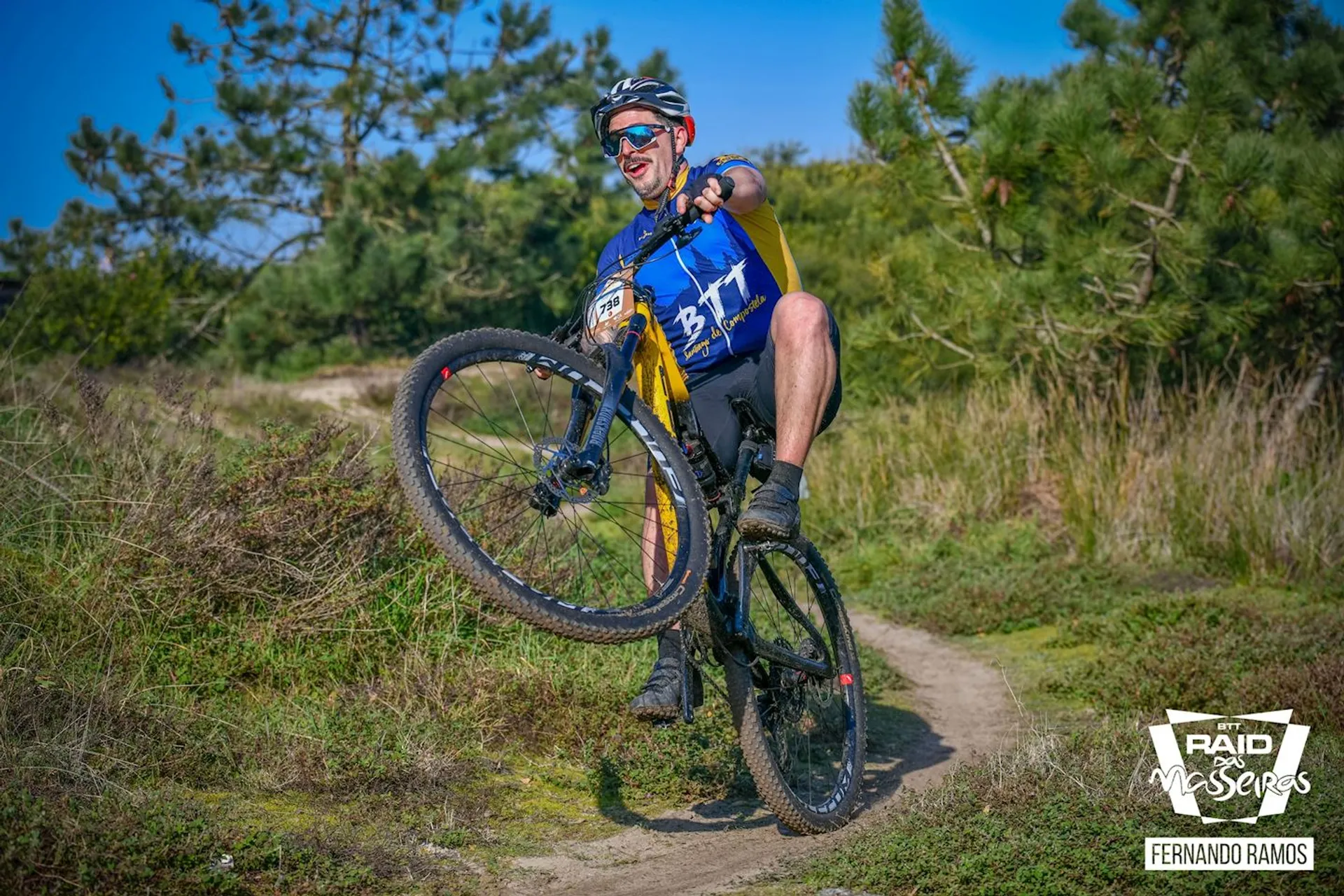 This image shows a person riding a mountain bike on a trail. The cyclist is wearing a helmet, sunglasses, gloves, and a sports jersey with a number bib, indicating they might be participating in a competitive event or race. The rider is lifting the front wheel of the bike, performing a wheelie, displaying skill and control. The setting is outdoors with grass and bushes in the background, suggesting a natural trail environment. The weather appears to be clear and sunny.