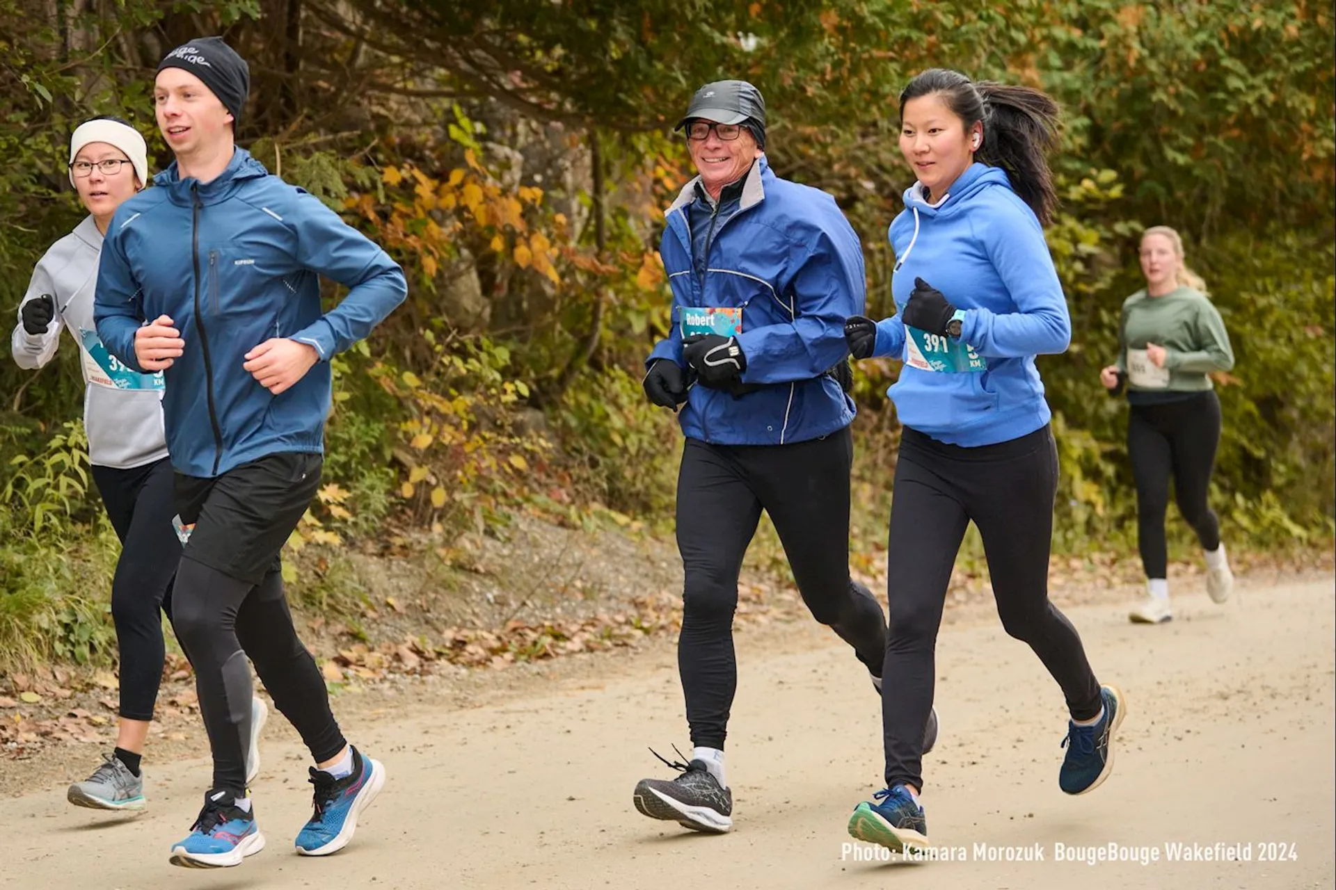 The image shows five people jogging or running on a dirt path through a wooded area. They are wearing athletic clothing suitable for cool weather. The scene appears to be part of a running event.