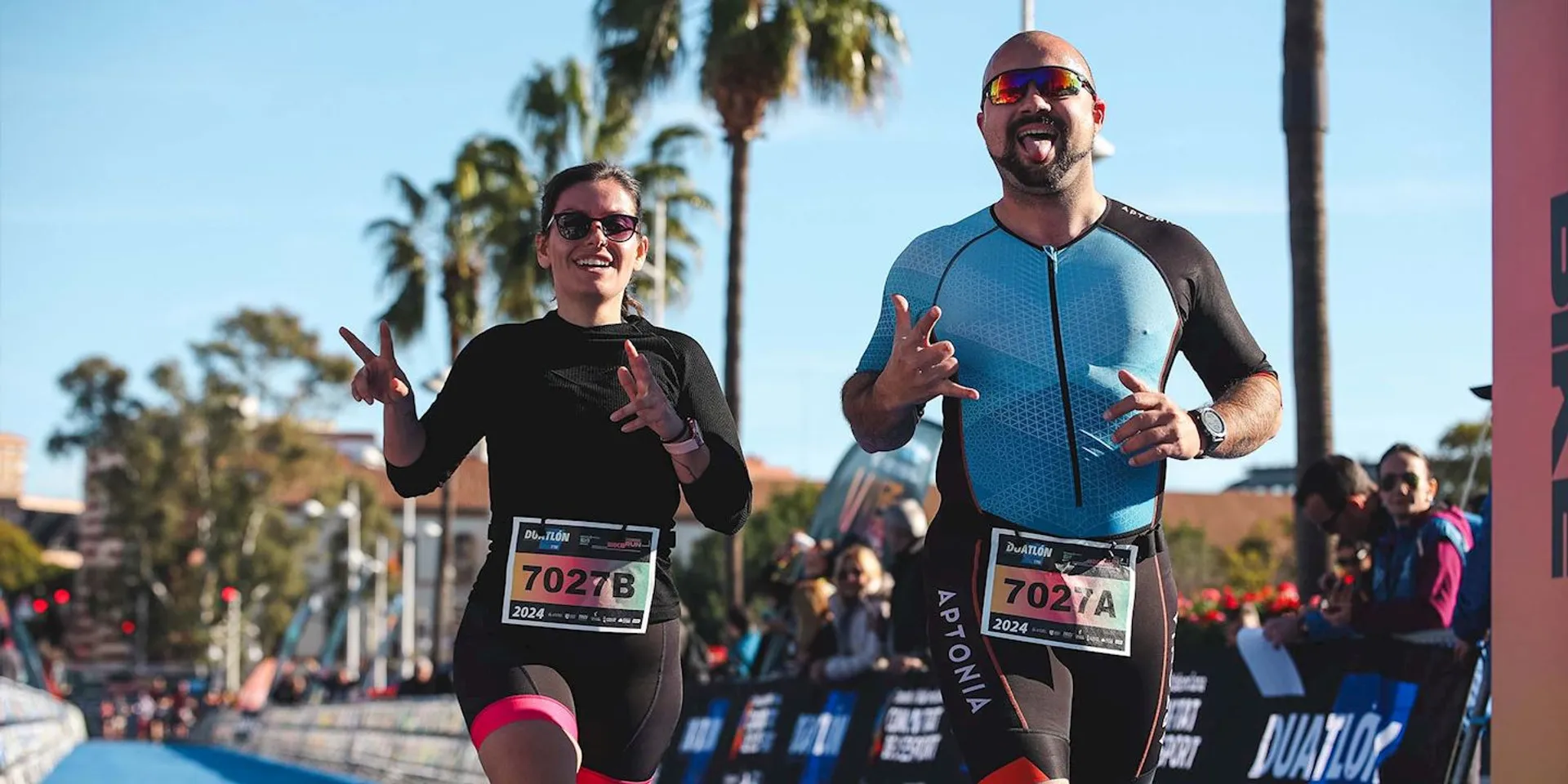 This image shows two participants in a running event, likely a marathon or a triathlon, as they appear to be wearing race bibs and the man is wearing what seems to be a tri-suit, which is common attire for triathlon participants. They are crossing the finish line, as indicated by the branded archway above them, smiling and gesturing with their hands. This suggests they are happy and possibly celebrating their achievement. It's a sunny day with clear skies, and there are spectators on the sides, which adds to the event's festive atmosphere.