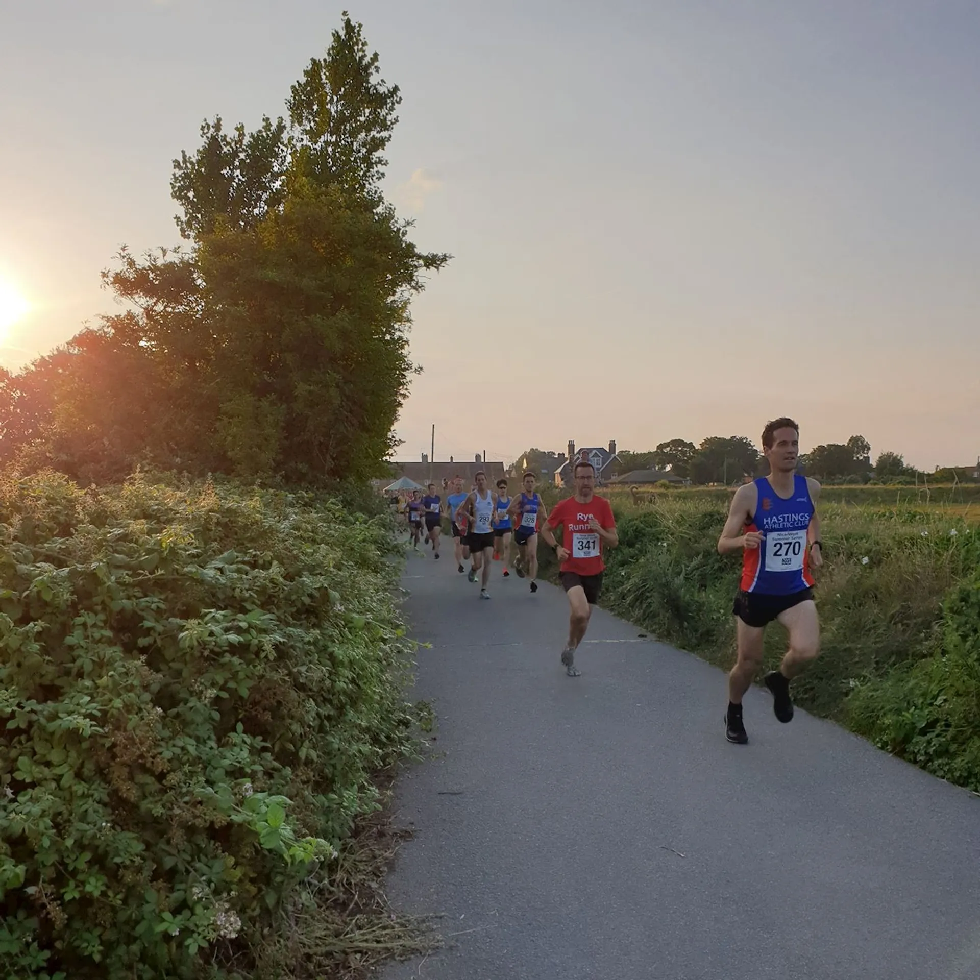 The image shows a group of people running on a paved path, likely participating in a race. They are wearing numbered bibs, and the setting appears to be outdoors with greenery and a clear sky at sunset.