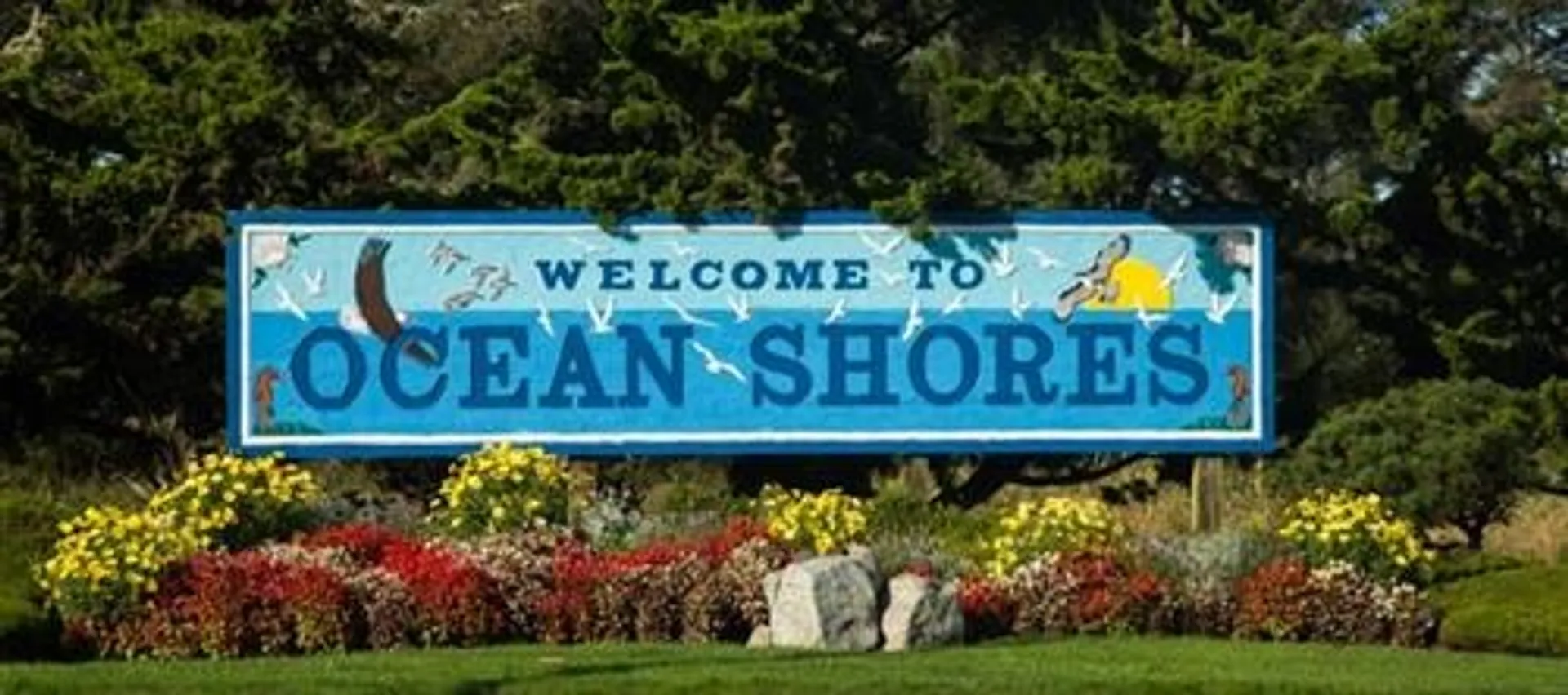 The image shows a large welcome sign that reads "Welcome to OCEAN SHORES" against a vibrant blue background with artistic depictions of marine life, such as fish and a whale's tail. Below the sign, there is a well-manicured garden area with a variety of colorful flowers and shrubbery. The setting appears to be a sunny day with some green trees in the distance, potentially indicating that the sign is located in a coastal or beach town area called Ocean Shores.