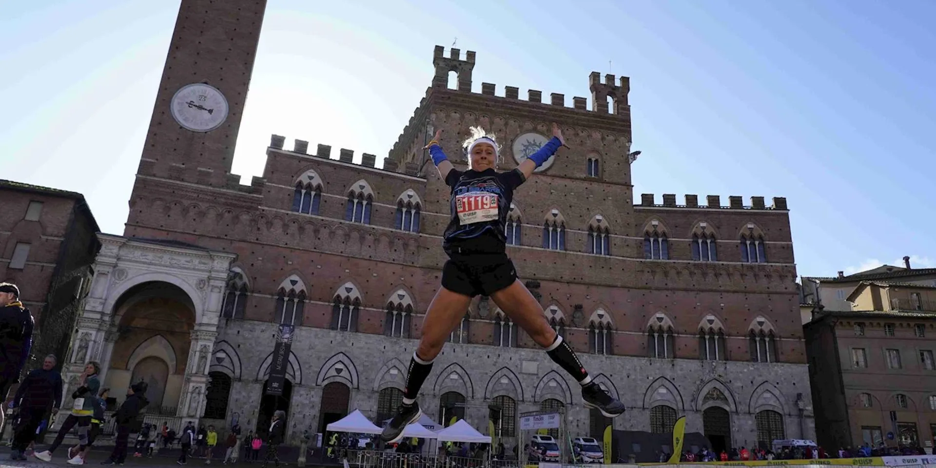 The image shows a joyful person mid-jump with their arms up and legs bent back, adding a sense of excitement or triumph. They seem to be celebrating or showing enthusiasm, possibly after a run or during a public event, based on the bib number indicating participation in a race. The background features an imposing historical building with crenellations and a tall clock tower, suggesting the location is a square in a city with rich architectural heritage. The blue sky suggests it's a nice day, and there are other people and tents around, indicating some type of outdoor event is taking place.