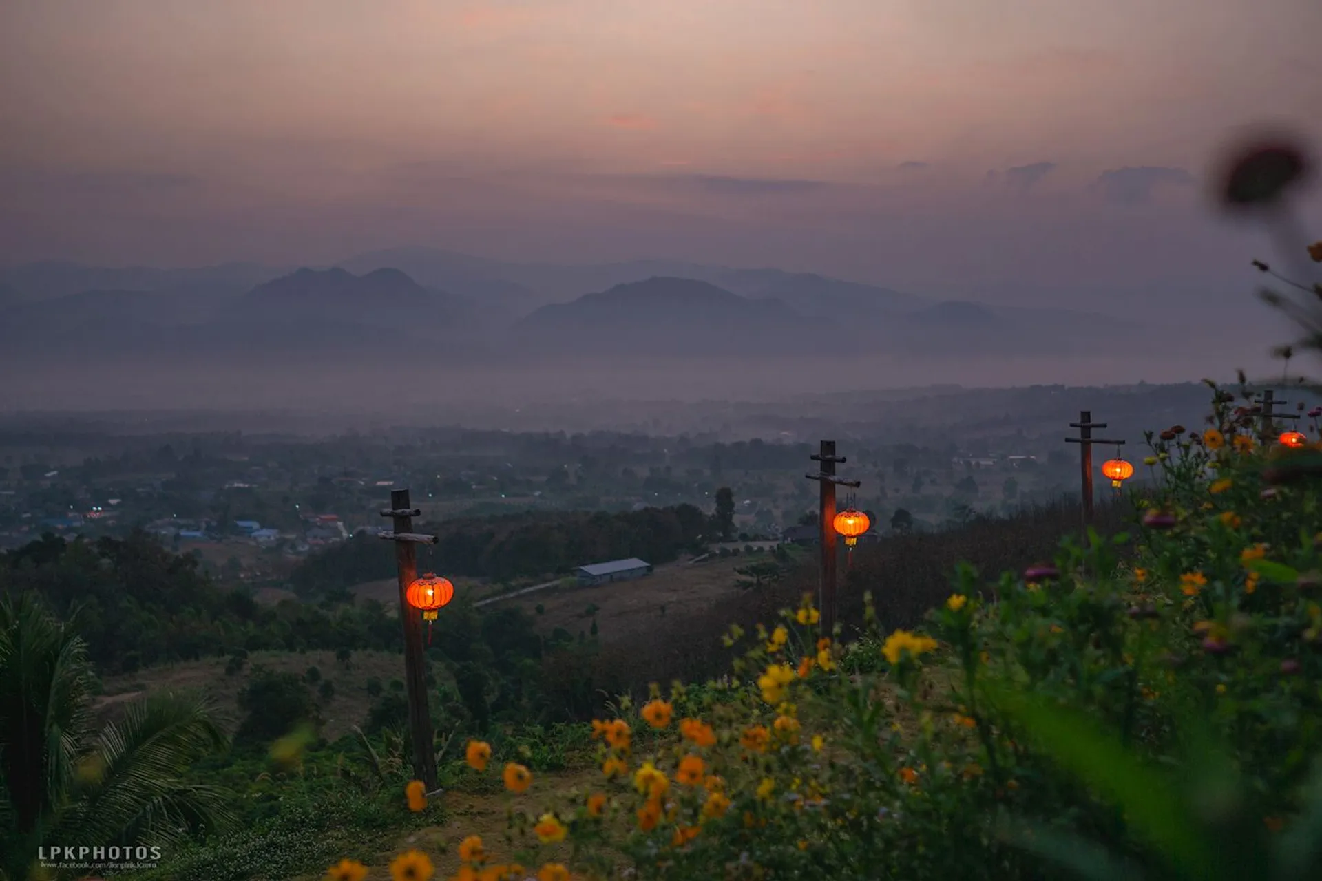 The image shows a scenic landscape at dusk or dawn. There are mountains in the background, with mist or fog creating a hazy effect. In the foreground, there are red lanterns and a variety of flowers. The scene conveys a peaceful and serene atmosphere, with soft lighting and natural beauty.