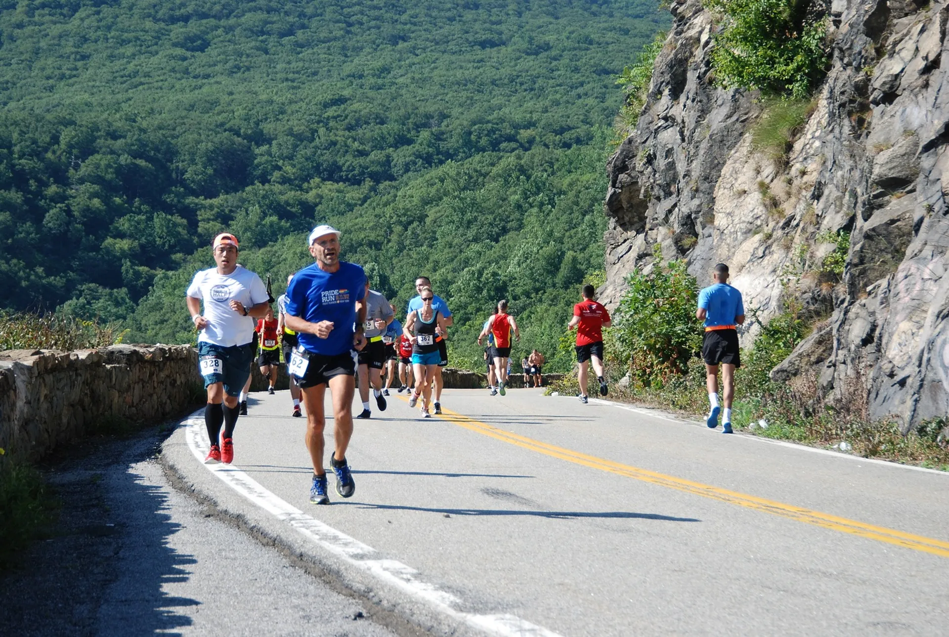 The image shows a group of runners participating in a road race. They are running on a paved road with a rock face on one side and what appears to be a hill or mountain region on the other. The runners are dressed in typical athletic attire suitable for running, such as shorts, t-shirts, and running shoes. Some appear to be wearing hats or visors, possibly to shield their eyes from the sun. The scenery suggests a sunny day, and the natural environment indicates that this race might be in a rural or mountainous setting. The runners are spread out along the road, each at their own pace.