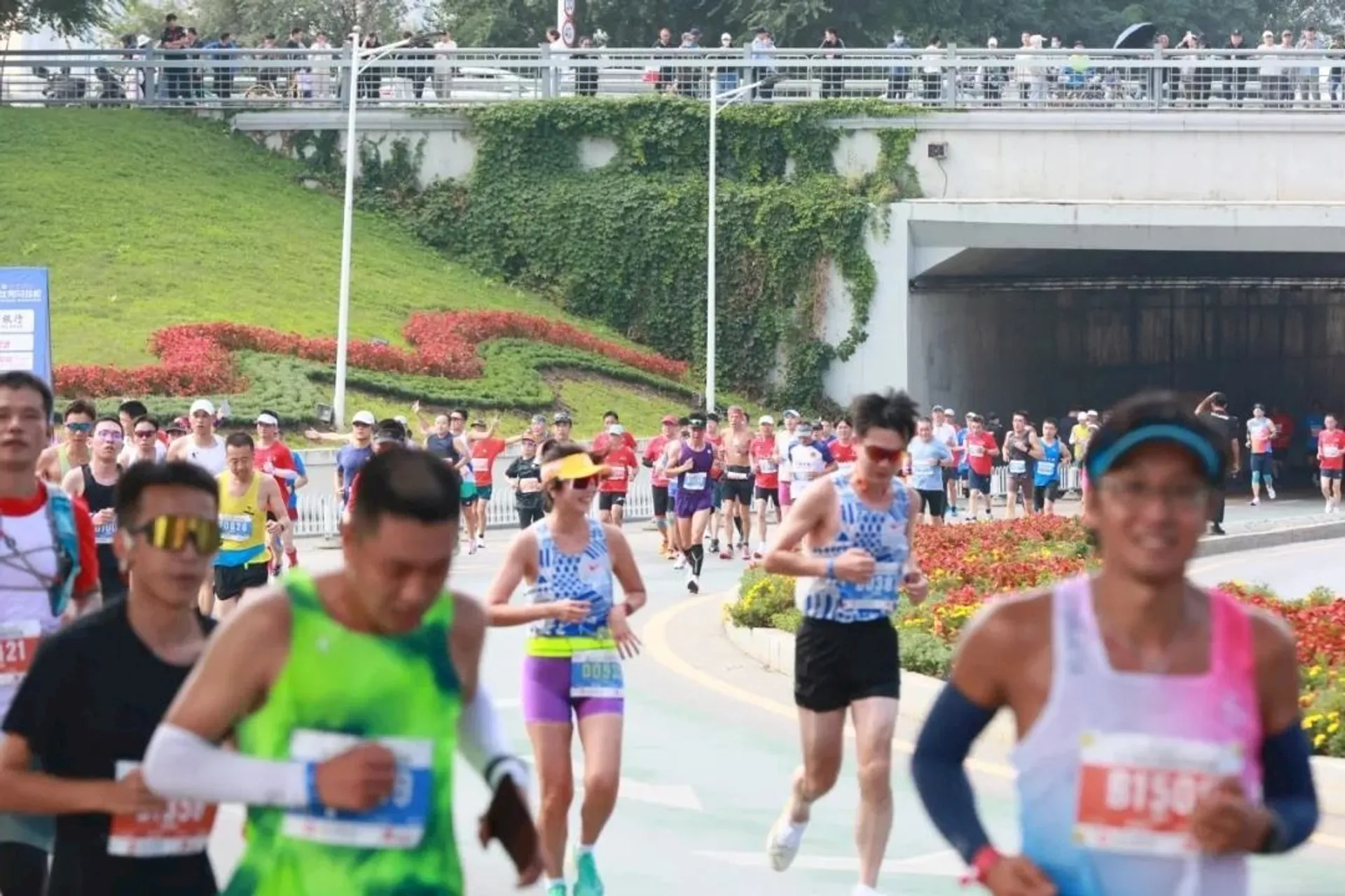 The image shows a group of people running in a marathon or organized race. The runners are on a road, wearing athletic gear with bib numbers. There are spectators and greenery in the background.