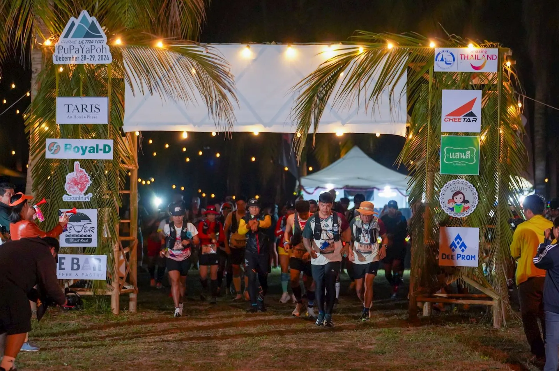 The image shows a group of people participating in a night-time running event. Runners are starting or passing through a banner marked with various logos and sponsors. It's dark, with palm trees and string lights in the background, creating a festive atmosphere.