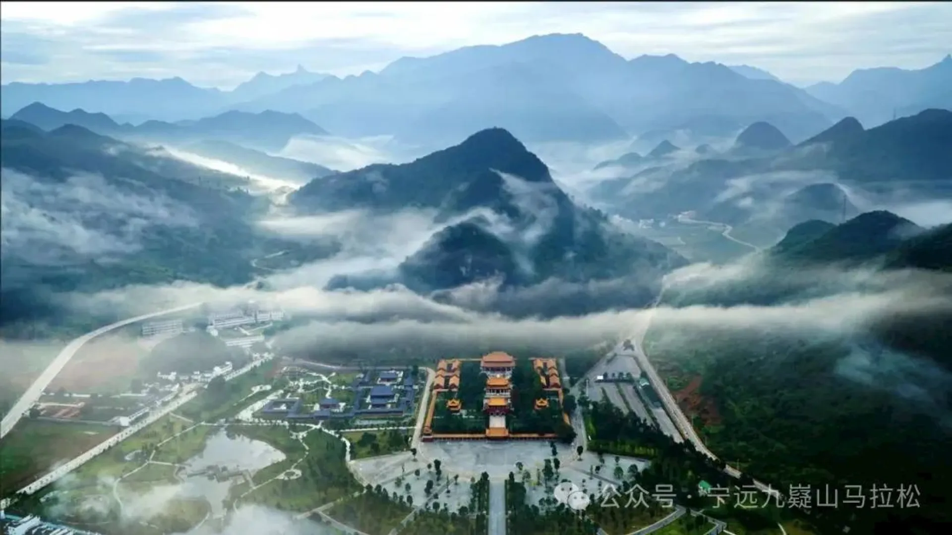 The image depicts an aerial view of a landscape featuring mountains and valleys partially covered in mist. In the center, there's a large architectural complex that resembles a traditional temple or palace with multiple structures and courtyards. The surrounding area includes greenery and possibly water features. The background shows a range of mountains under a clear sky.