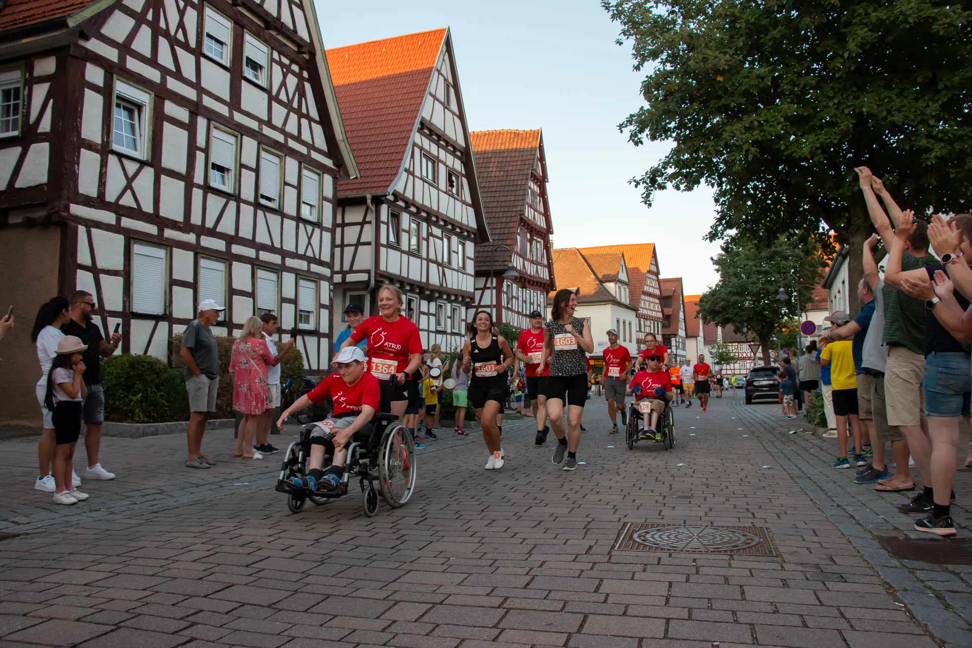 In the image, we see a group of people participating in what appears to be a running event or marathon. There are a few individuals in wheelchairs who are being pushed by runners, suggesting this event may be inclusive, supporting participants with disabilities. Spectators on both sides of the road are clapping and cheering on the participants, creating a supportive and encouraging atmosphere. The road is lined with traditional half-timbered houses, indicating this may be taking place in a European town, possibly Germany due to the architecture style. It's a scene of community involvement and physical activity, likely meant to promote health, inclusion, and togetherness.