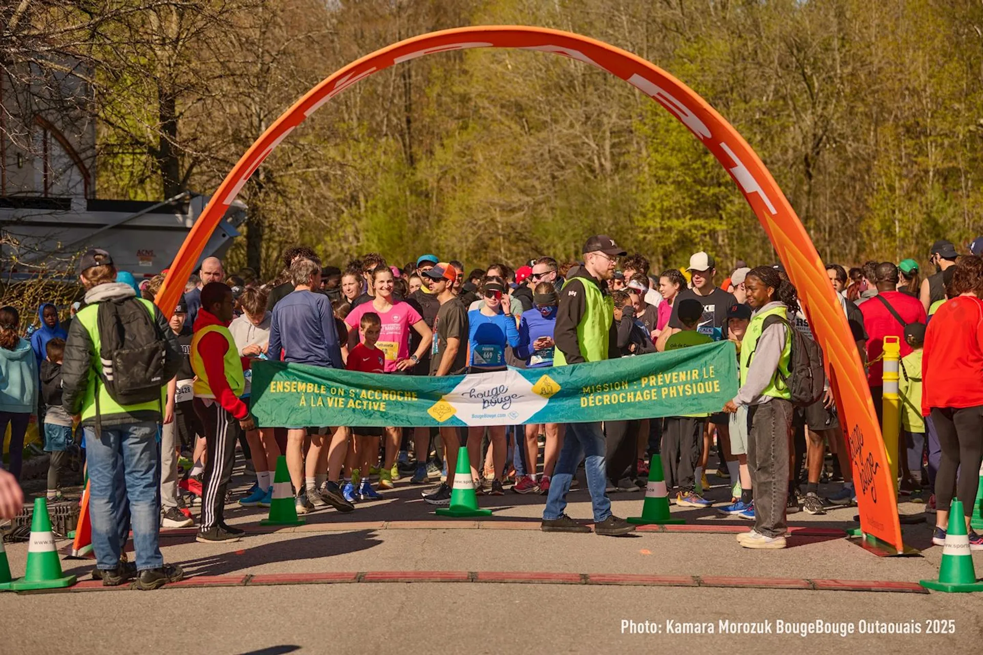The image shows a group of people gathered at the starting line of a race. There is an orange arch and several participants are wearing running attire. Some individuals are holding a banner with text on it, and there are volunteers in safety vests. The event seems to have a festive atmosphere.