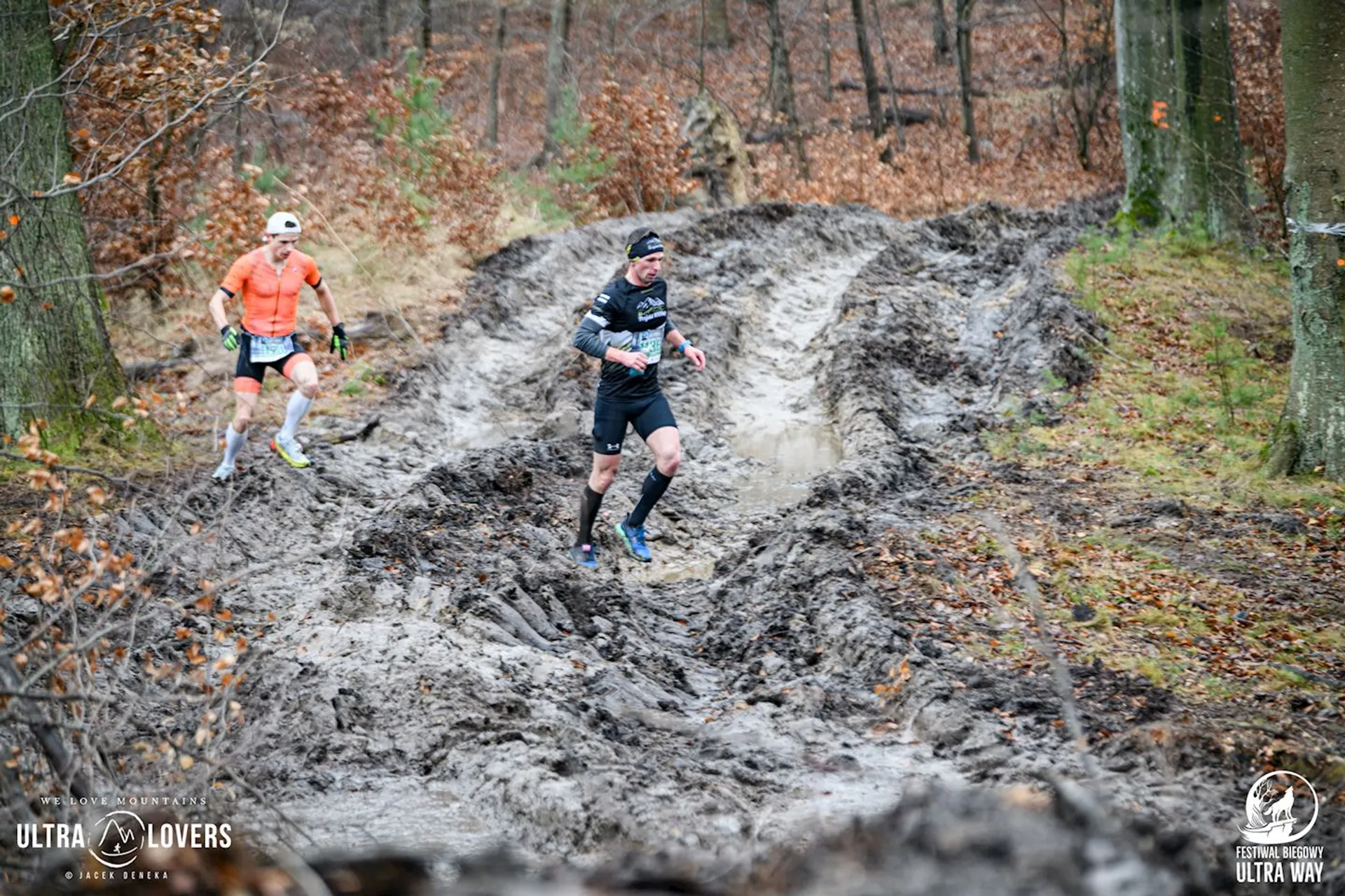The image shows two people running through a muddy and rough terrain in a forested area. They're wearing athletic gear, suggesting they're participating in a trail run or race. The area around them is filled with leafless trees and brown foliage, typical of early spring or late fall. There are logos in the corners indicating it's an event, possibly related to an ultramarathon or trail running.