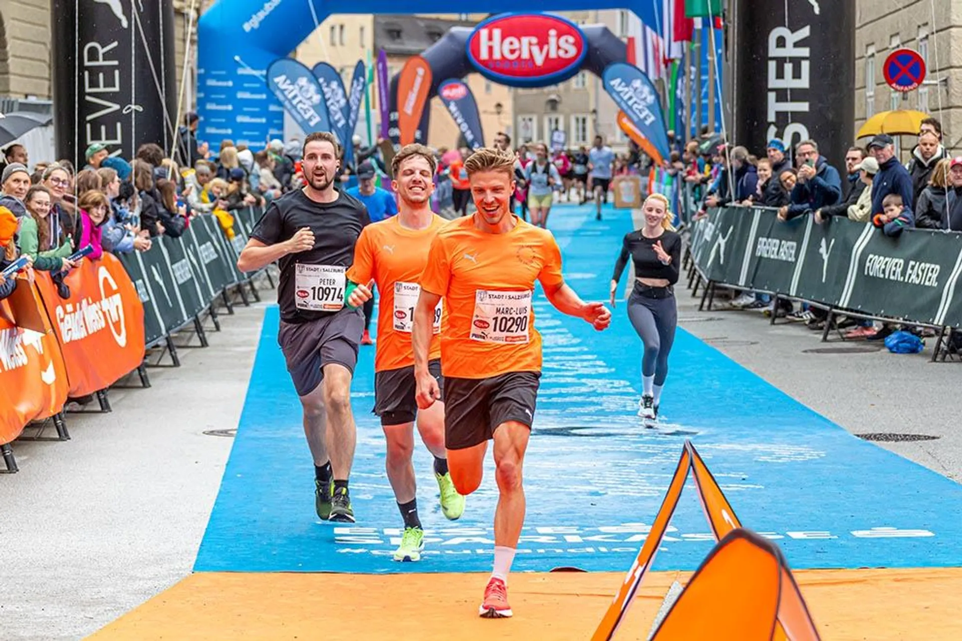 The image shows a group of runners participating in a race. The scene captures runners crossing the finish line on a blue carpet, surrounded by cheering spectators. In the background, there are banners and signs, some with logos and numbers. The atmosphere looks energetic and celebratory.