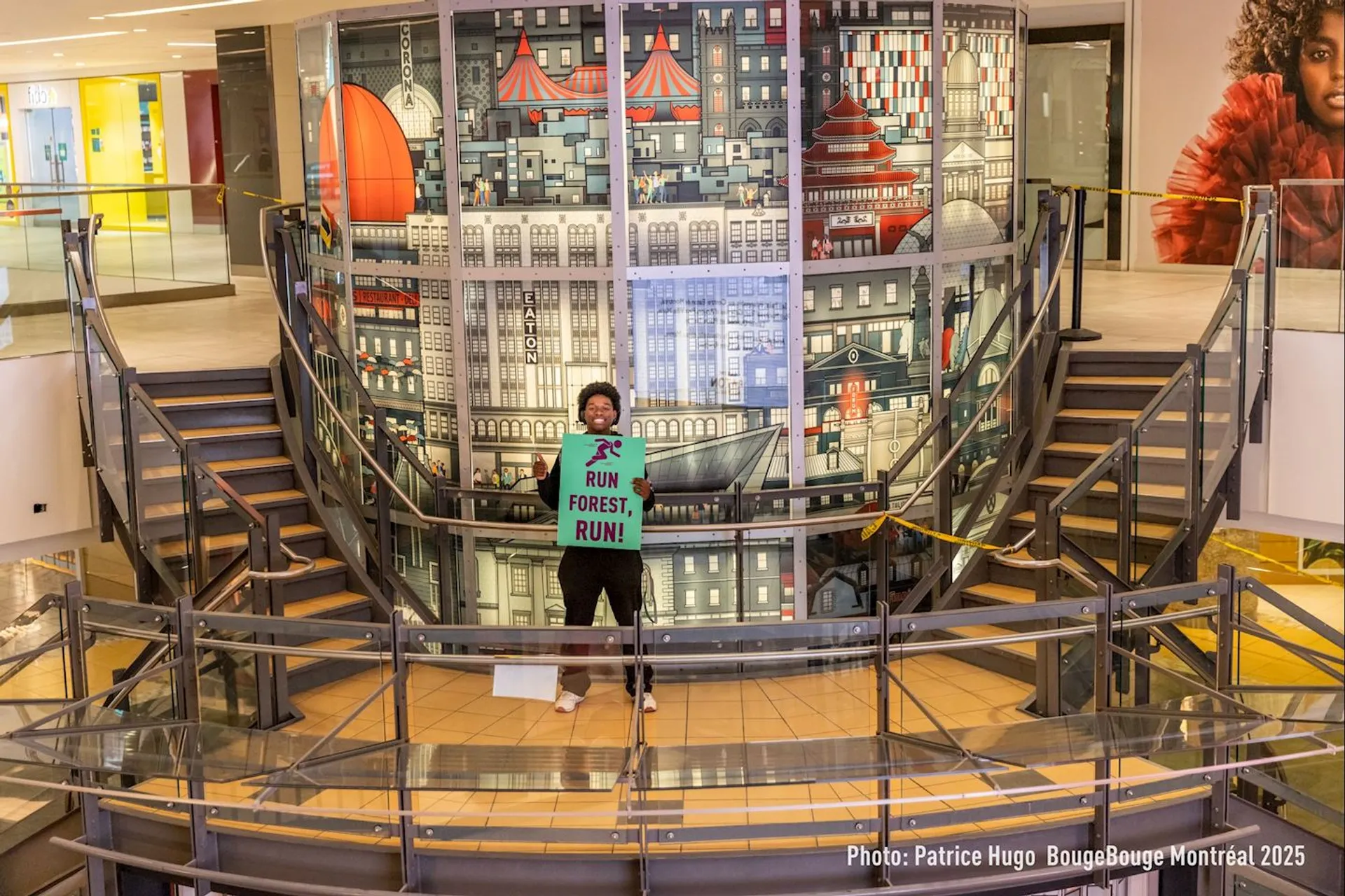 The image shows a person holding a sign that says "RUN FOREST, RUN!" They are standing in a building with a decorative wall behind them featuring a cityscape design. The area appears to be an indoor space with railings and a staircase.