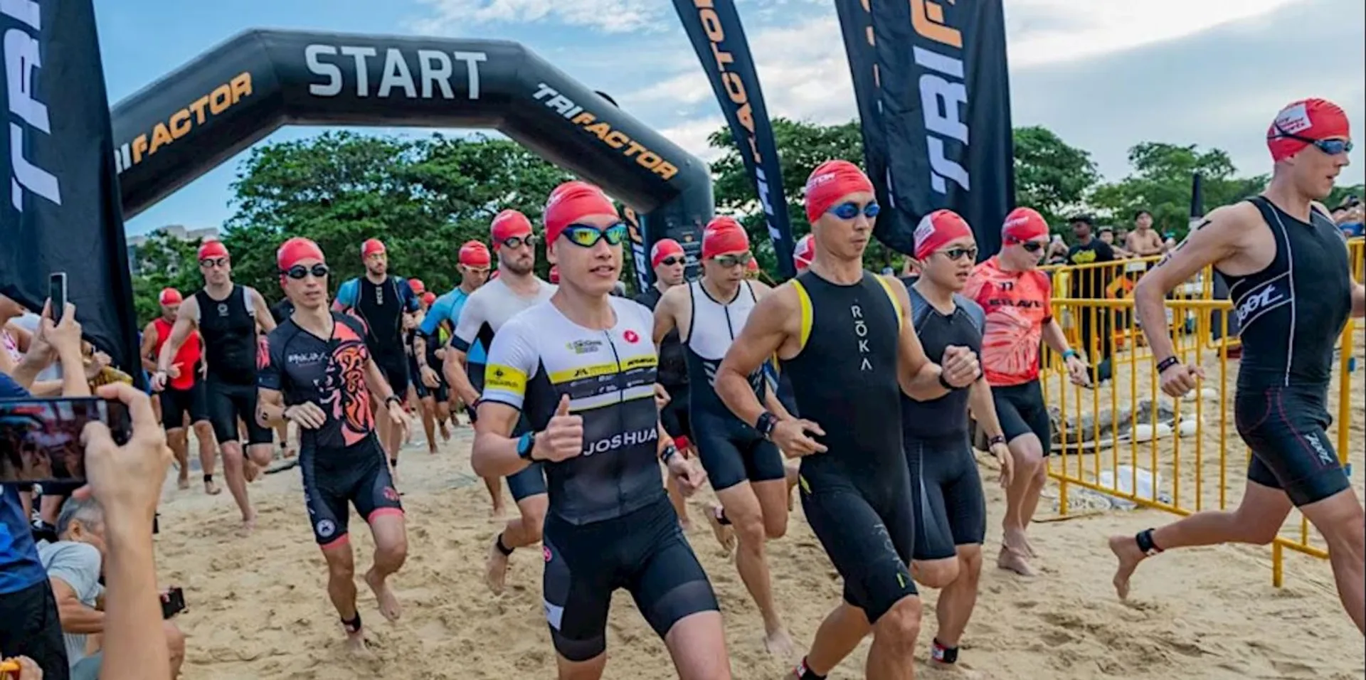 The image shows a group of athletes participating in a triathlon event. They are wearing swimsuits and red swim caps, indicating they may be starting the swimming portion of the event. There are banners and a start line gate with "TRI" visible on them. The event appears to be taking place on a beach.