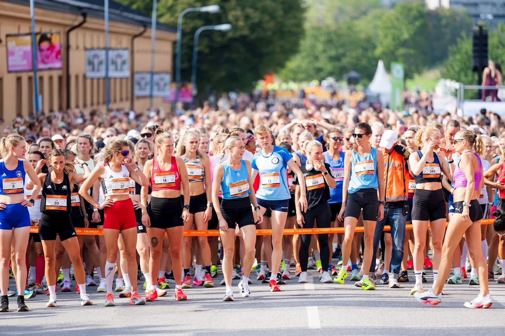 The image shows a large group of runners gathered at the starting line of a race. The participants are wearing athletic clothing, race bibs, and are ready to start running. It's a sunny day, and there are trees and buildings in the background.