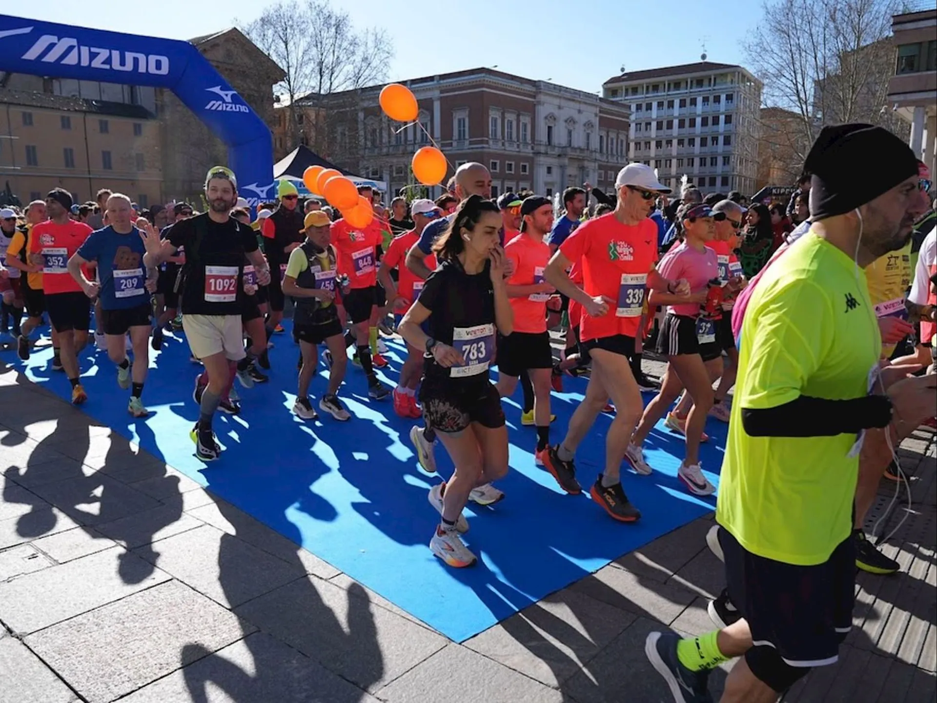 This image shows a group of people participating in a running event or race. They are running under a Mizuno-branded arch. Various participants are wearing numbered bibs and athletic clothing, and some orange balloons are visible in the crowd. The event takes place outdoors in a city area with buildings in the background.