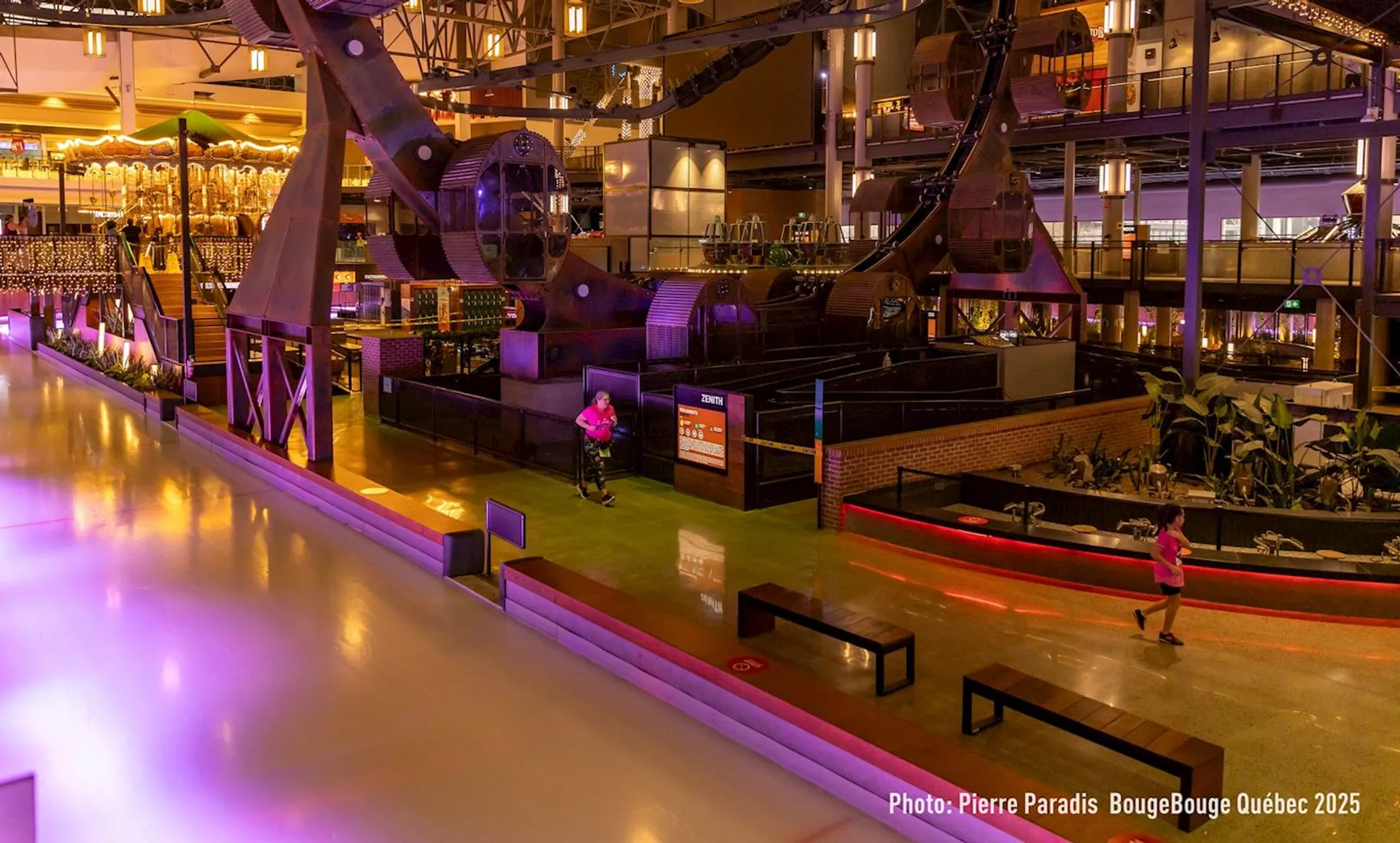 The image shows an indoor space with an ice-skating rink and a modern, industrial-style decor. There are colorful lights, benches, and a carousel in the background. A few people are visible near the rink and walking around.