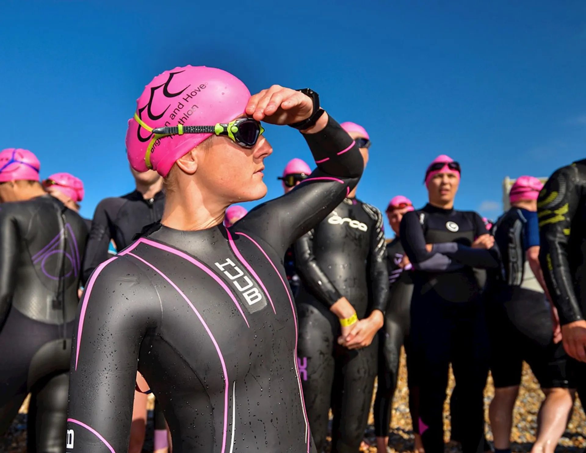The image shows a group of people wearing wetsuits and bright pink swim caps, likely preparing for a swimming event or triathlon. One person in the foreground is adjusting their goggles and looking towards the distance. The setting appears to be outdoors, possibly on a beach or near a body of water.