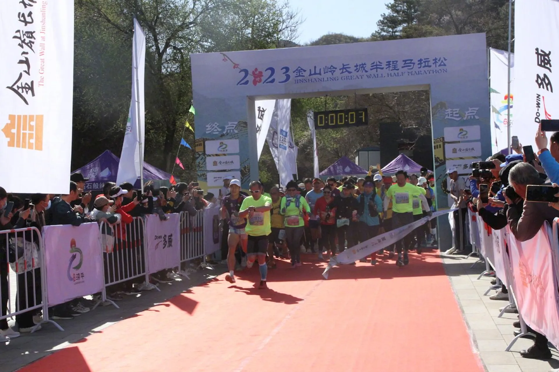 The image shows a group of runners crossing the starting line of a half-marathon race. They are just beginning the race, as the digital clock above the start/finish gate displays "00:00:01," indicating one second has passed since the start. This is the 23rd edition of the Jinshanling Great Wall Half Marathon, as indicated by the banner. Spectators are lining both sides of the starting gate, watching and some taking photos of the event. The race appears to be organized and festive, with banners and flags around, and it seems like a bright, sunny day.
