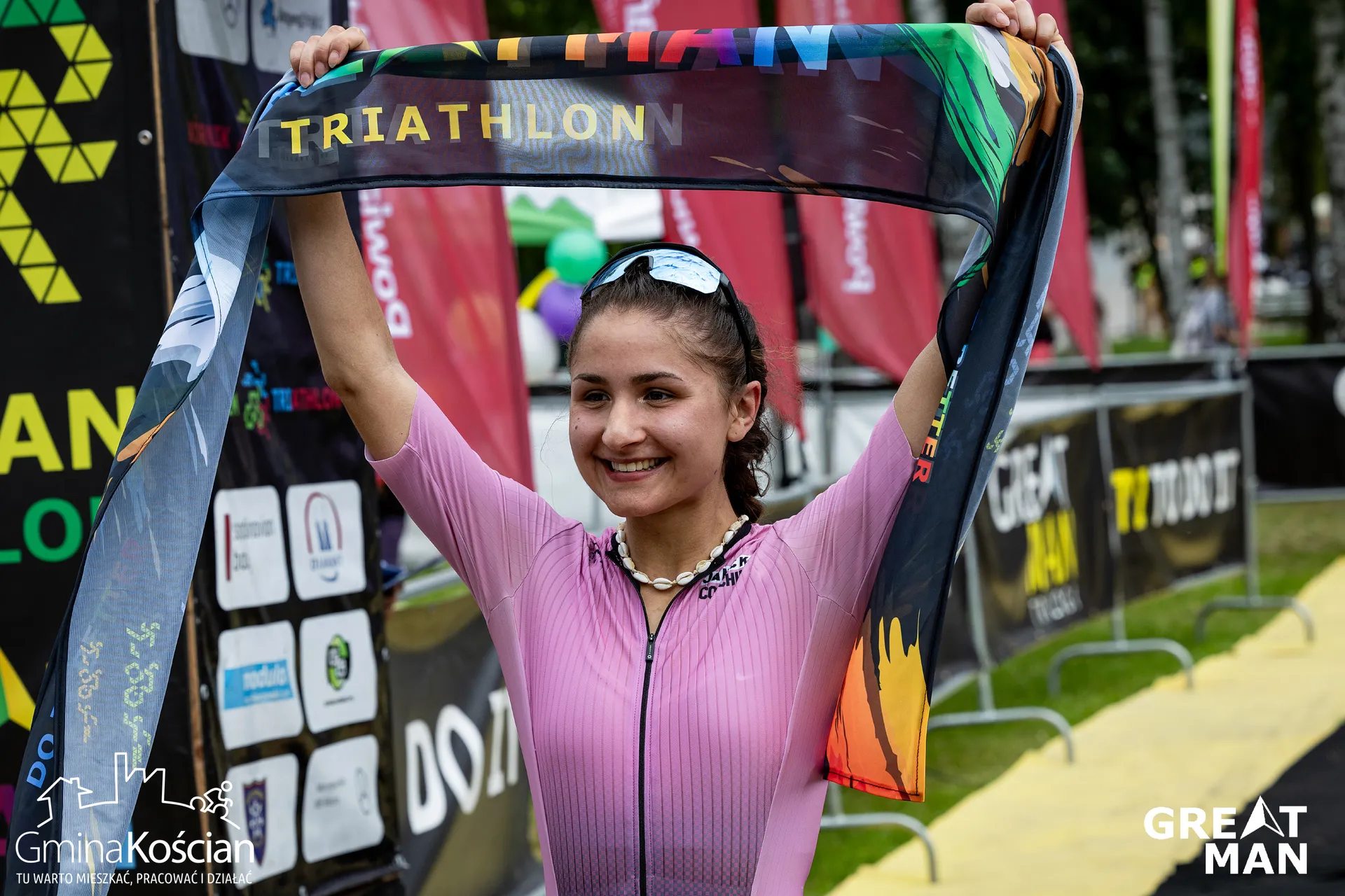 The image shows a person at a triathlon event holding a banner overhead. They appear to be celebrating or finishing a race. There are sponsor logos and event signs visible in the background.