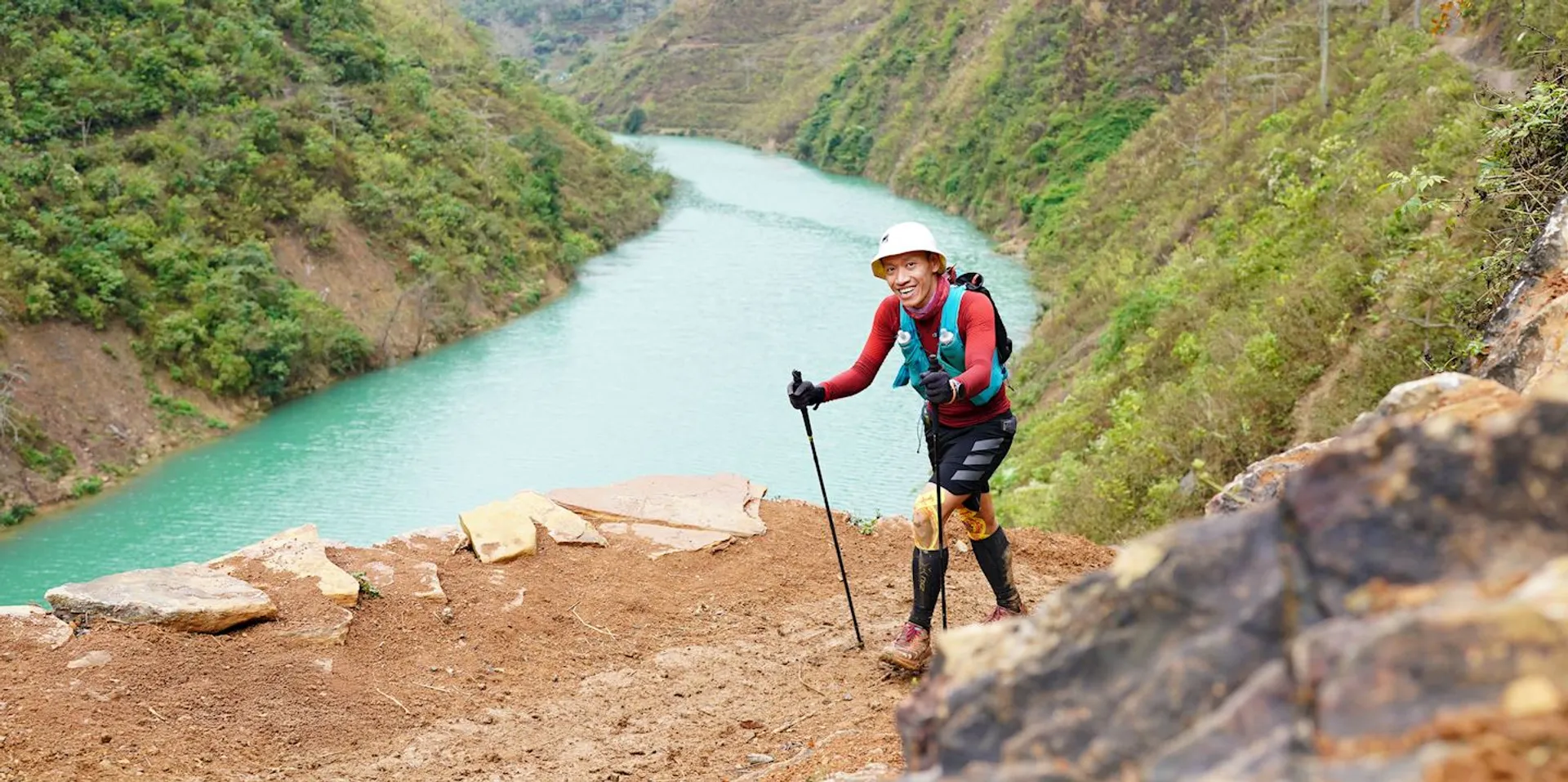 The image shows a person hiking on a trail beside a river. The person is using trekking poles and wearing a helmet and outdoor gear. The landscape includes a turquoise river running between green, hilly terrain.