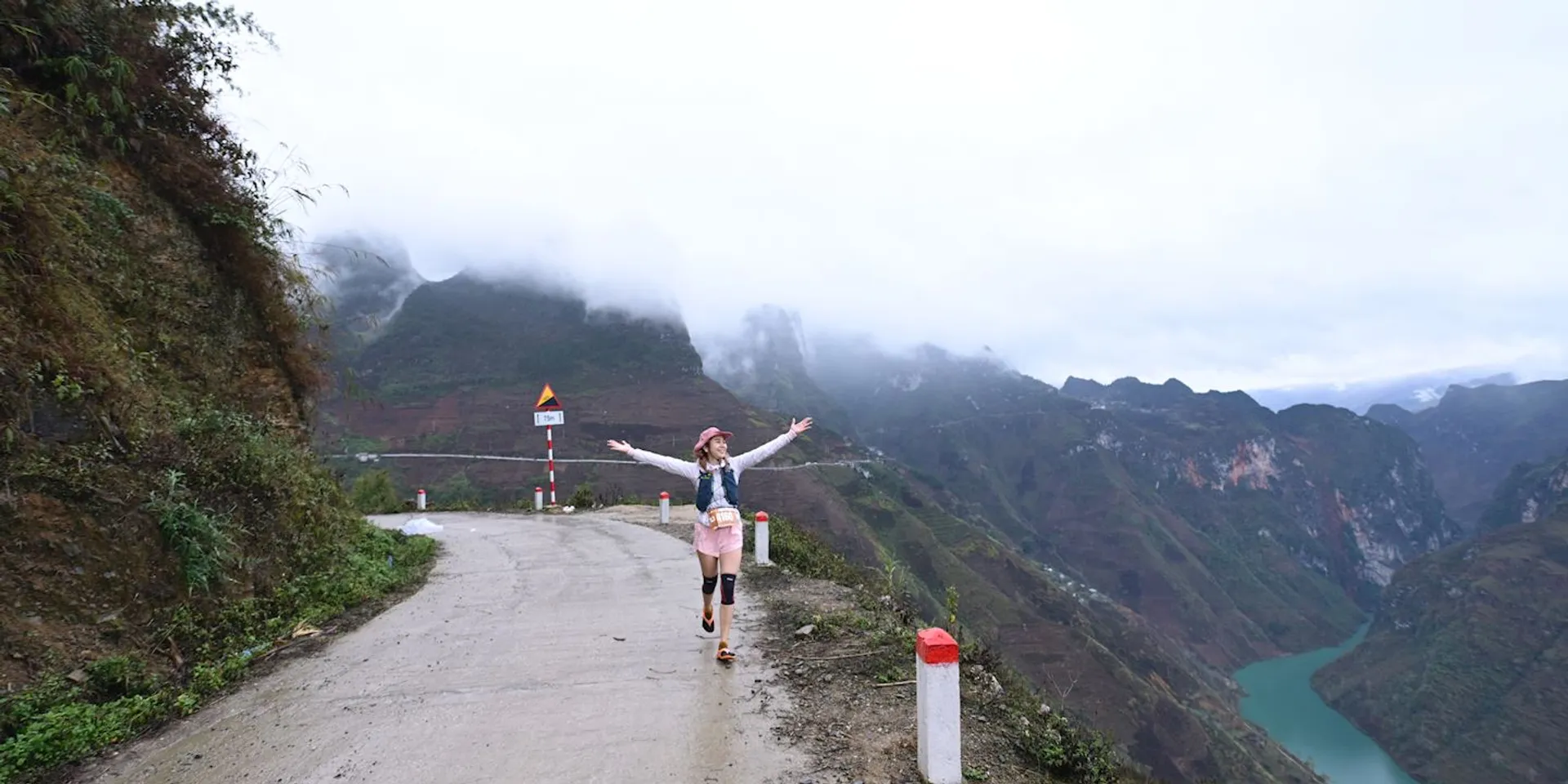 The image shows a person joyfully walking on a mountain road with arms outstretched. The scene includes lush green mountains, a winding river, and a misty sky, suggesting an atmosphere of freedom and adventure. The road is bordered by white and red poles, and the background features dramatic mountainous terrain.