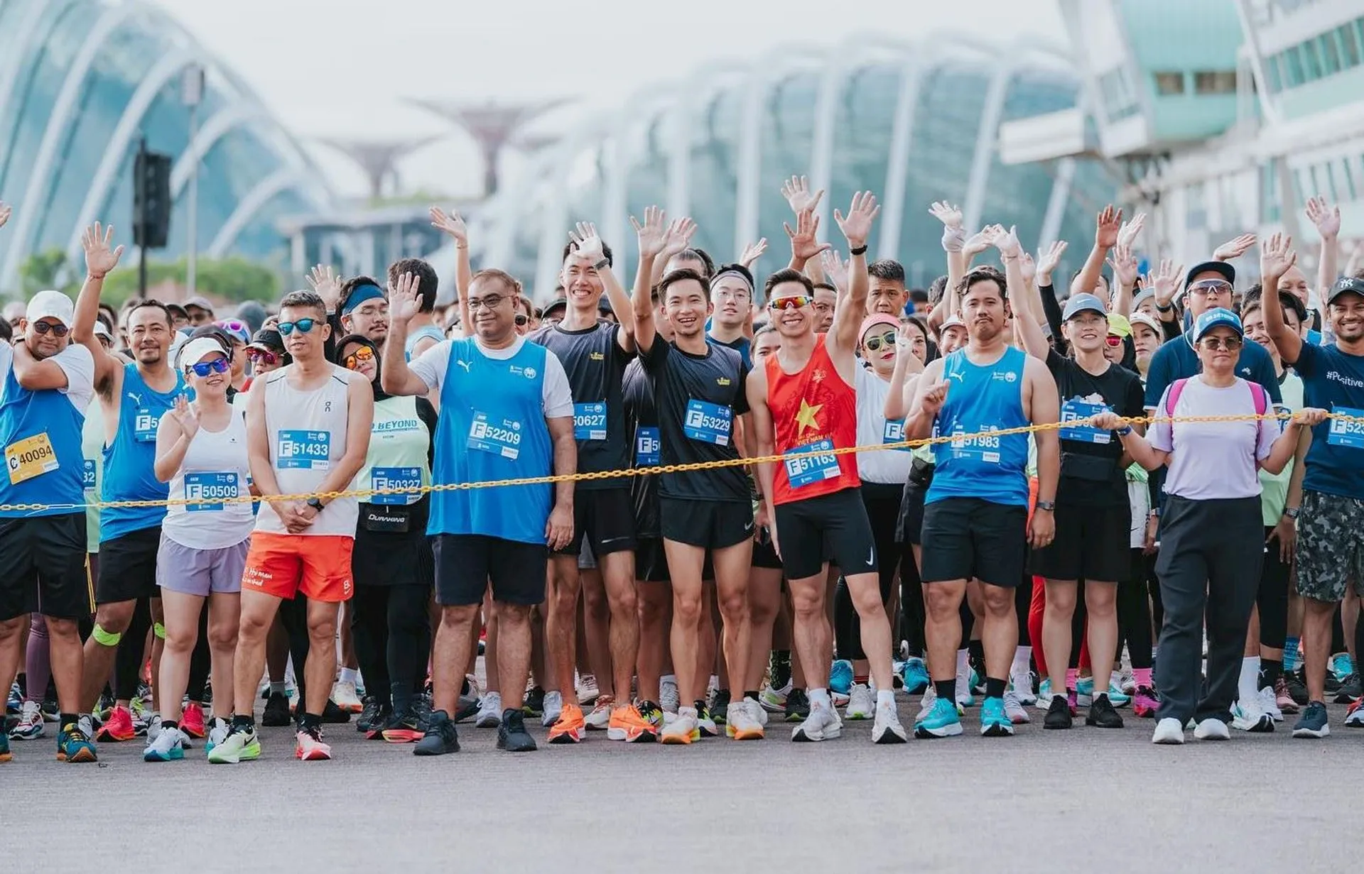 This image shows a group of people gathered for a running event. They are standing behind a starting line, dressed in athletic wear, and some are waving. The background features modern architectural structures.