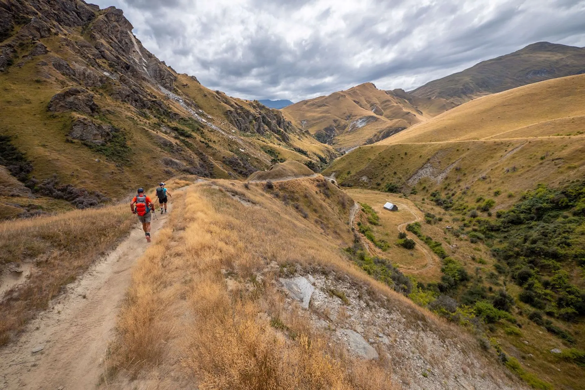 The image depicts a scenic outdoor trail with two individuals hiking. They are walking along