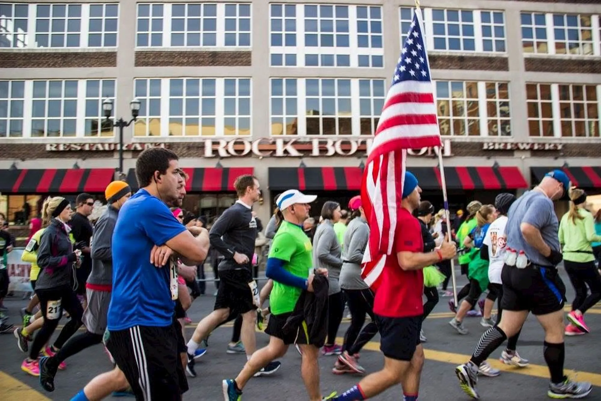 This image shows a group of people participating in a marathon or running event. They are running past a building with red and black awnings. One participant is carrying a large American flag.