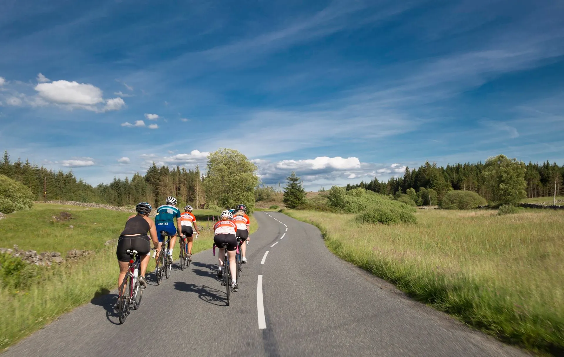 The image shows a group of cyclists riding along a road. The road appears to be in a rural or natural setting with open fields, trees, and a forested area in the background. The sky is mostly clear with some clouds, suggesting good weather conditions for cycling. The cyclists are wearing biking attire, which typically includes helmets and form-fitting cycling clothing. They seem to be in motion, as indicated by their postures and the position of their legs and feet on the pedals.