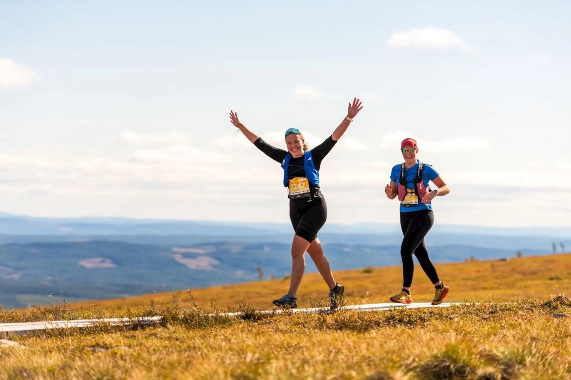 The image shows two people participating in an outdoor trail run or marathon on a wooden pathway in an open, natural landscape. One person is raising their arms joyfully, and both are wearing running gear and race numbers. The background features a vast horizon with hills or mountains under a clear sky.