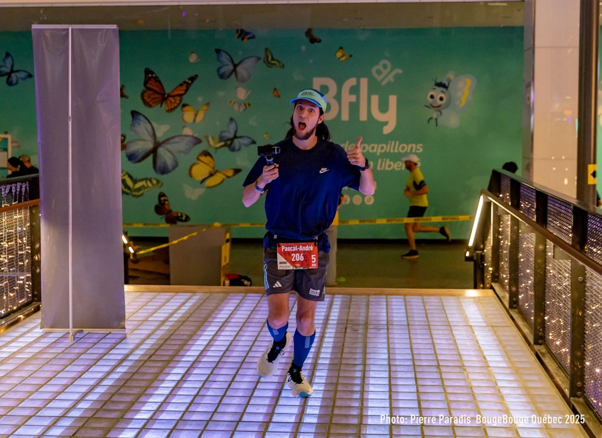 The image shows a person running indoors, likely part of a race or event. They are wearing athletic clothing, including a hat and sport shoes, and holding an object in their hand. The background features a colorful wall with butterflies and text related to an exhibit or event. The ground is lit with a glowing pattern. There's a banner with numbers on the runner suggesting their participant number. 