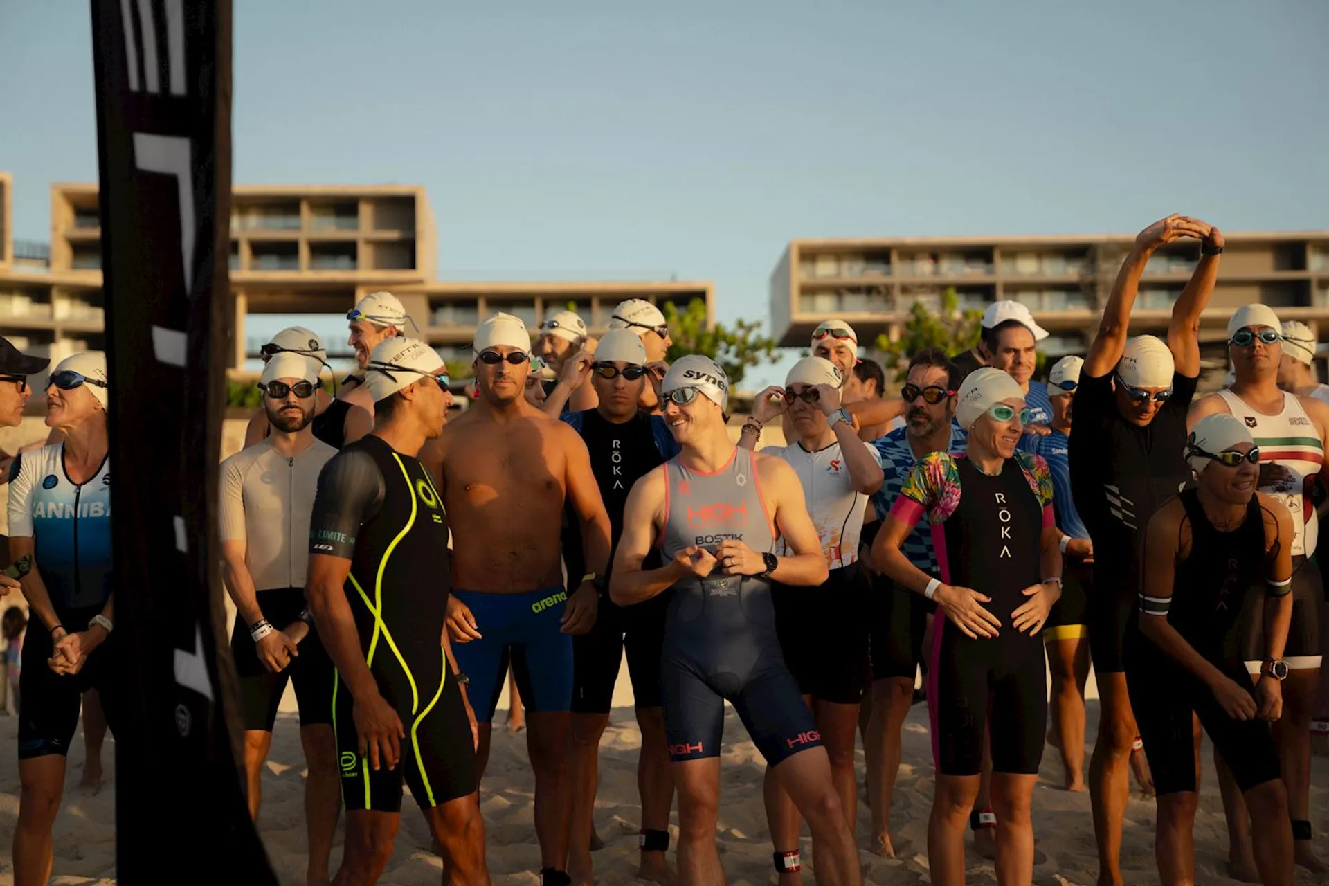 The image shows a group of people in swim gear, including swimsuits and swim caps, gathered together on a beach. They appear to be preparing for a swimming event or race, with some participants wearing goggles. The background features several large buildings.