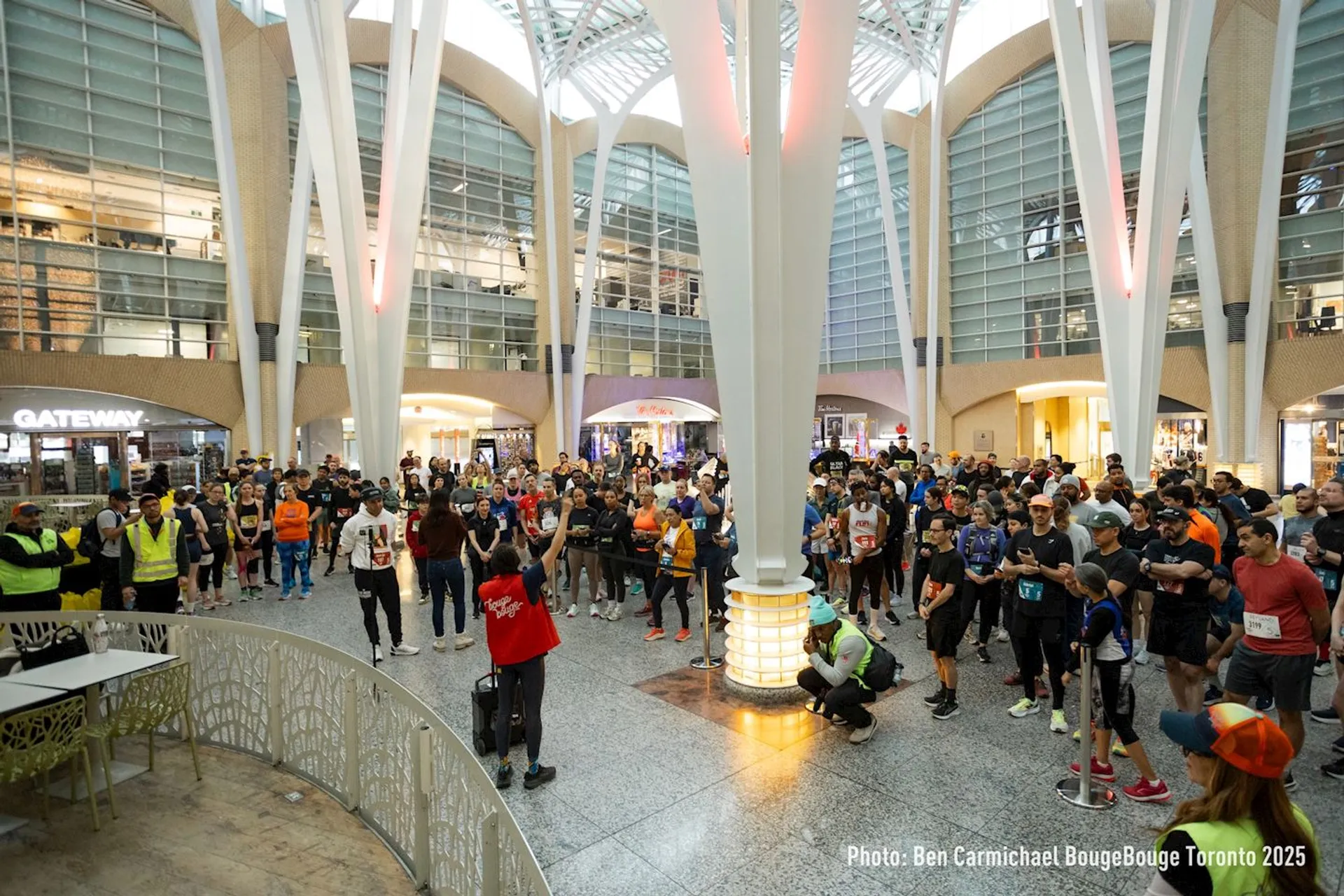 The image shows a group of people gathered inside a large, modern indoor space with high ceilings and white architectural columns. There are some people wearing running gear and numbers, suggesting an organized event, likely a race or fitness gathering. There's a person in front who appears to be speaking or addressing the crowd. The setting looks like a public or commercial area, and some onlookers are standing around.