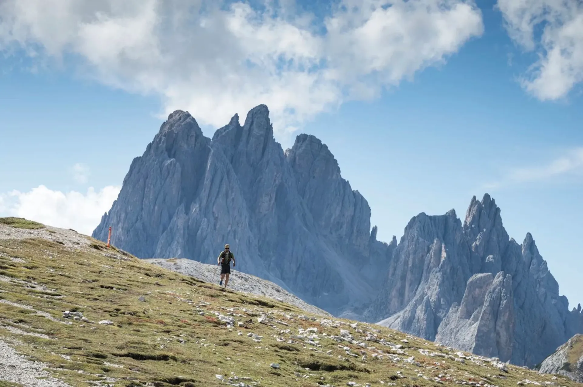The image shows a hiker walking on a grassy and rocky mountain slope. In the background, there are large, jagged mountain peaks under a partly cloudy sky. The scenery suggests a mountainous or alpine environment.