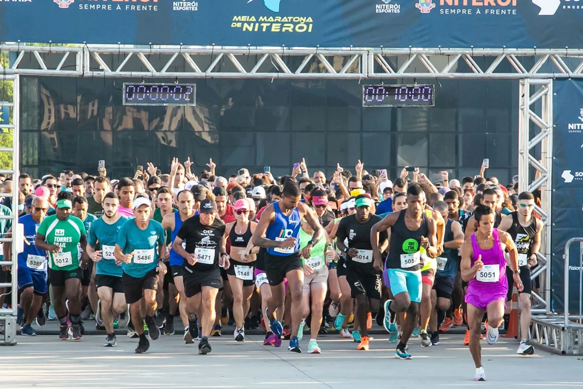 The image shows a group of runners at the start of a race. They seem to be just crossing the starting line, as we can observe a digital timer overhead displaying 00:00:02, indicating the race has just begun. Participants are wearing typical running attire, including shorts, t-shirts, tank tops, and running shoes, with many wearing race bibs displaying their participant numbers. The scene suggests a well-organized event, such as a half marathon, 10K, or 5K, given the presence of large start/finish structure, timing equipment, and sponsorship banners. The atmosphere is energetic, with the runners appearing eager and focused as they embark on their race.