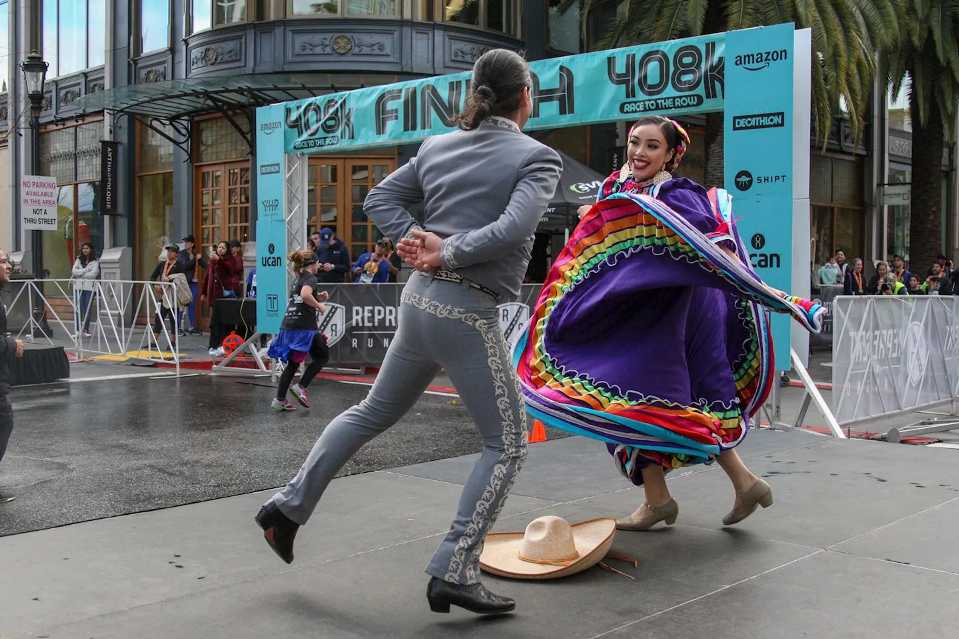 The image shows a pair of dancers performing a traditional dance in front of a finish line at an outdoor event. The female dancer is wearing a colorful, flowing dress, while the male dancer is dressed in a suit with a wide-brimmed hat on the ground. There are onlookers behind barriers, and the event appears to be called "408k," as seen on the banner above. The scene is lively and festive.
