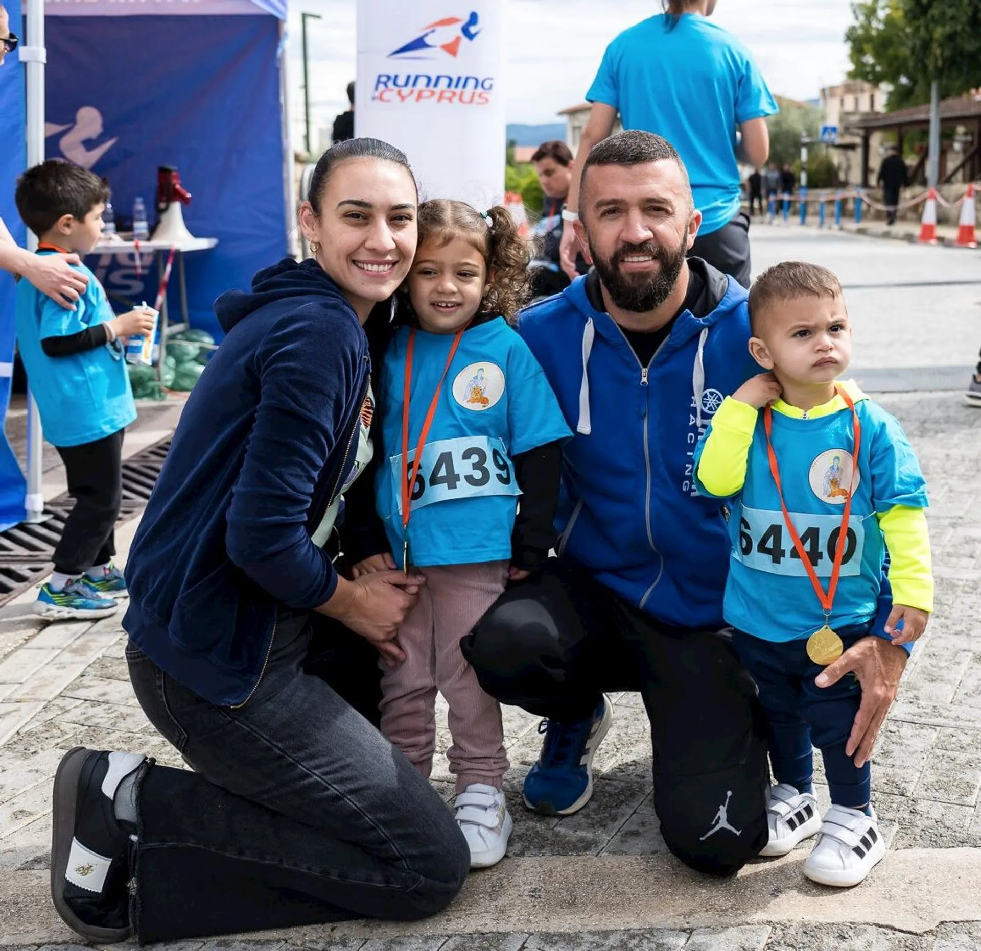 The image shows four people. There are two adults and two children, all wearing athletic attire. The children are wearing medals, and everyone appears to be smiling. They are posing together, likely at a running event, as suggested by the banner in the background and their race numbers.