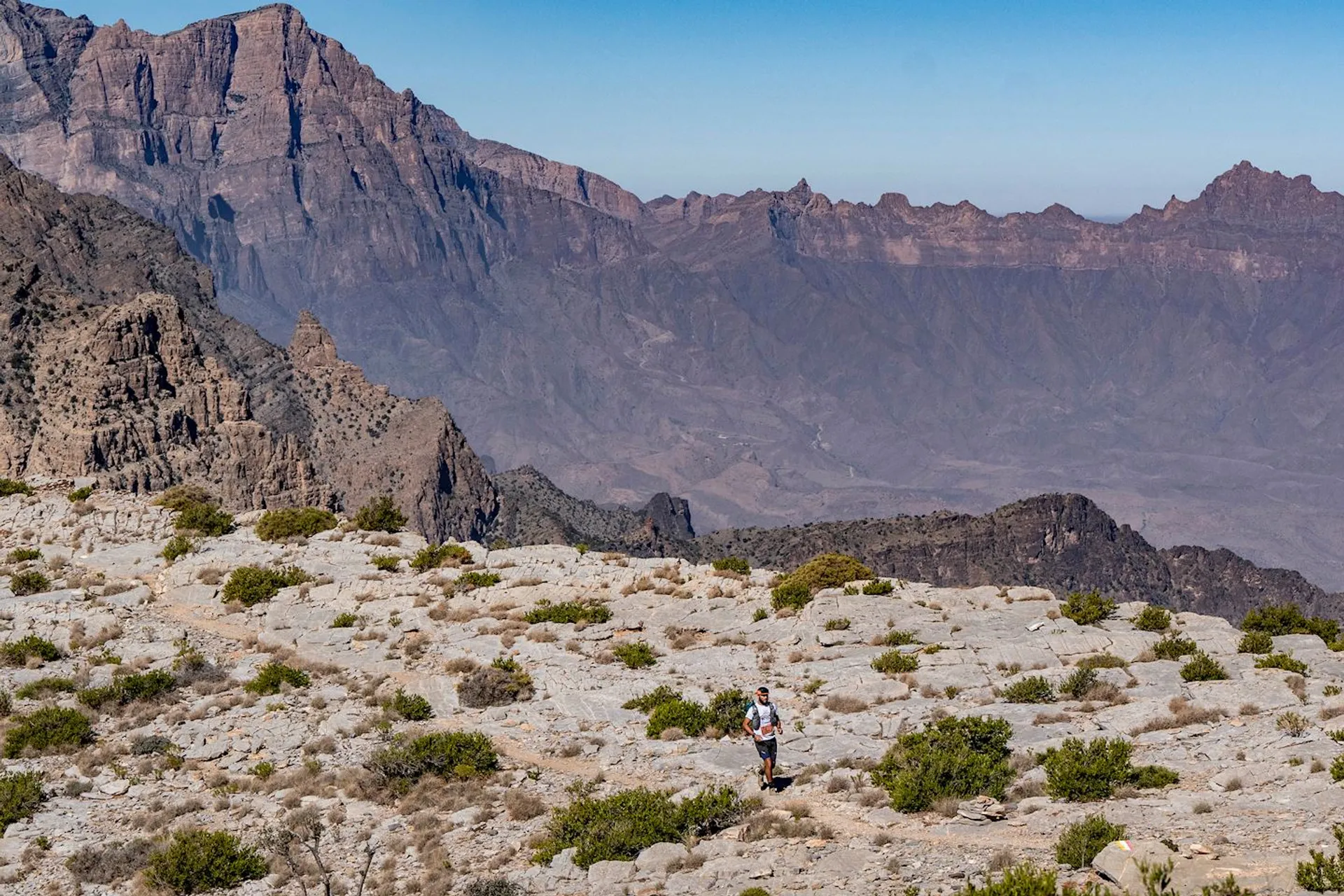 The image shows a rugged mountainous landscape with rocky terrain and sparse vegetation. A person is hiking in the foreground, and in the background, there are tall, jagged mountains under a clear blue sky. The scene presents a sense of vastness and natural beauty, ideal for outdoor activities like hiking or trekking.