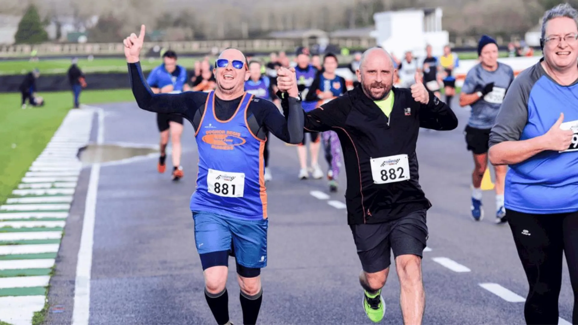 The image shows a group of people running a race. Two men in the foreground, wearing numbered bibs 881 and 882, are holding hands and appearing triumphant. They are surrounded by other runners, and everyone seems to be participating in an organized running event. The atmosphere is energetic and positive.
