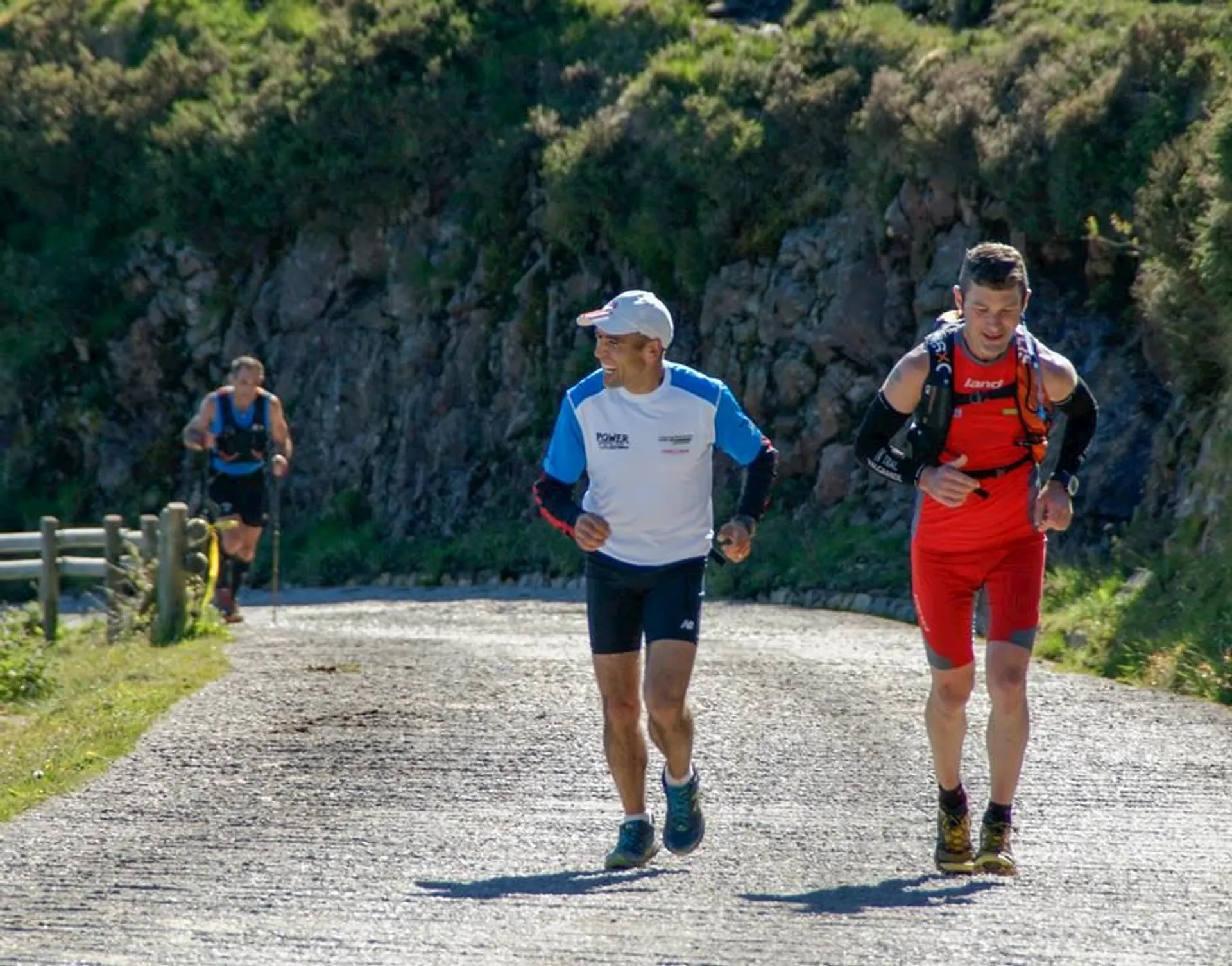 The image shows three people running on a road in an outdoor setting. Two of them are in the foreground, with one wearing a red outfit and a hydration pack, while the other wears a white and blue shirt and a hat. There is a third person running in the background. The environment looks natural, with greenery and rocky terrain.