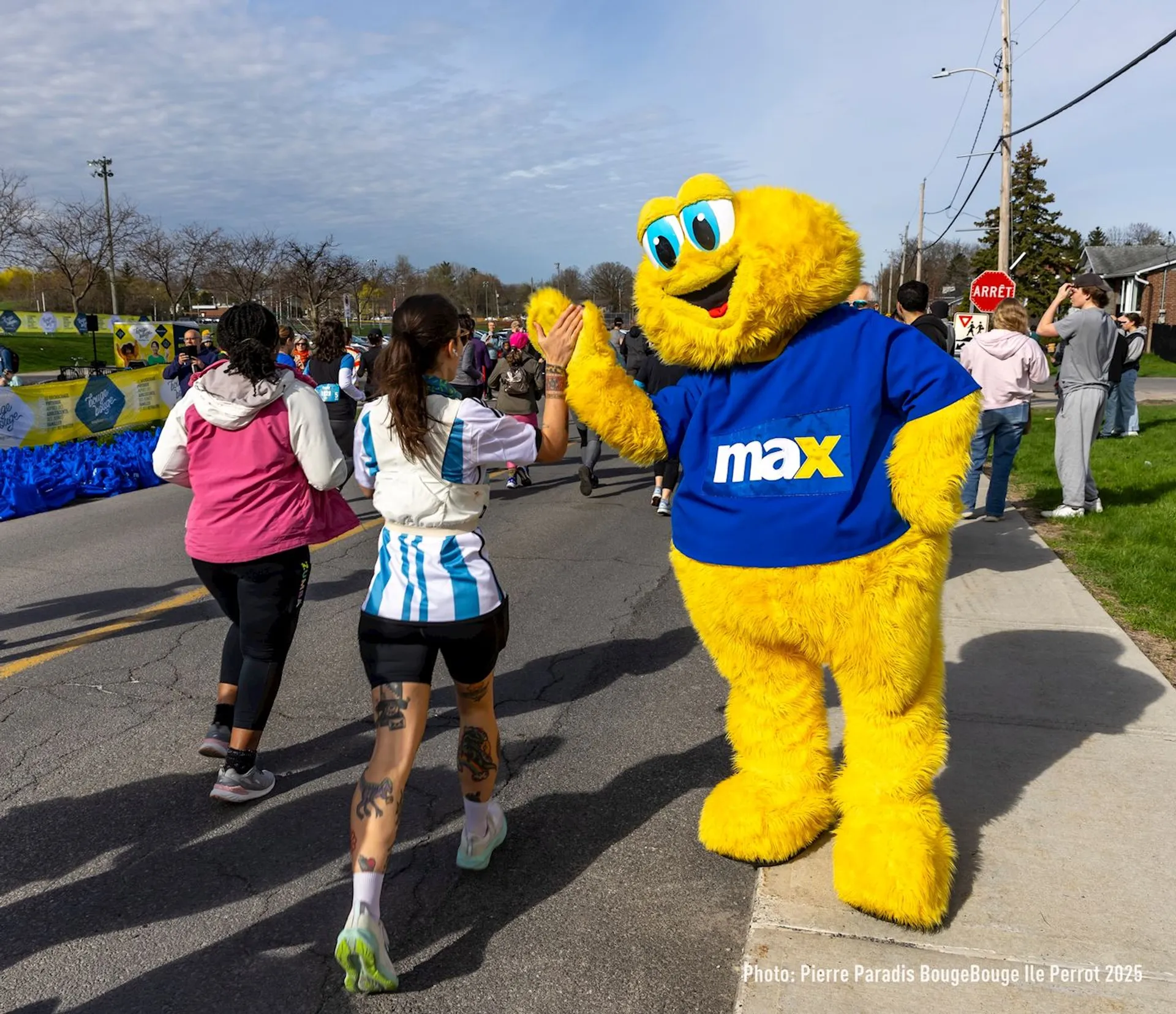 The image shows a person in a yellow furry costume, possibly a mascot, wearing a blue shirt with "max" written on it. The mascot is giving a high-five to a runner during an outdoor event, likely a race. There are other runners and spectators in the background.