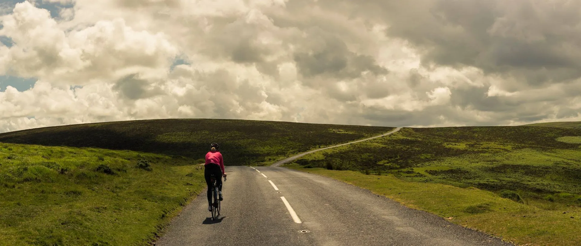 The image features a person cycling on an open road that cuts through a rolling landscape. The cyclist is wearing a bright pink top which provides a nice contrast to the natural greenery of the grassy hills around. The sky is filled with large, fluffy clouds, suggesting a day with good weather. The open road ahead curves slightly, indicating a vast and tranquil rural area, likely to be peaceful and ideal for a leisurely bike ride.