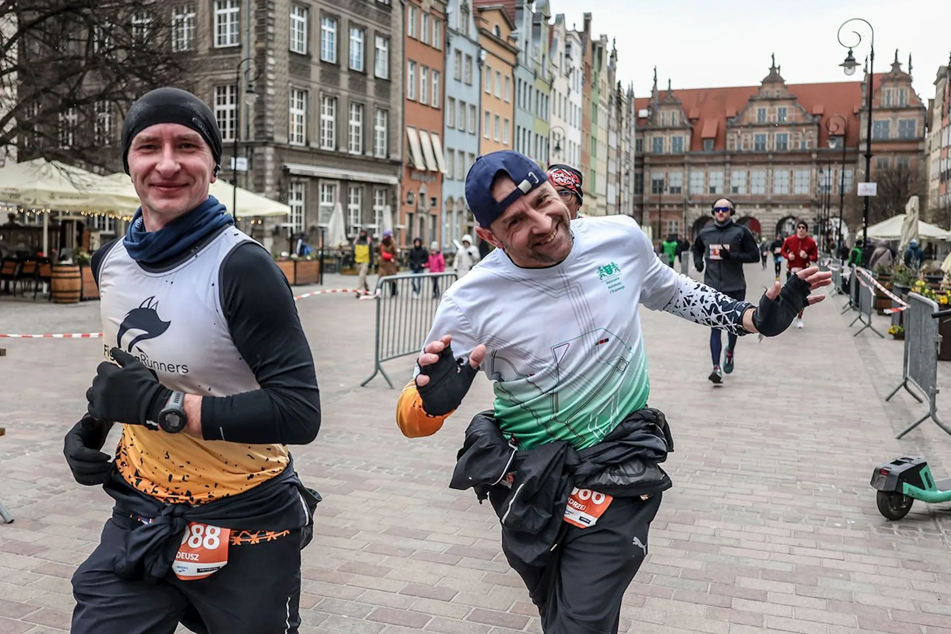 The image shows two runners participating in a race on a street lined with colorful buildings. They appear to be enjoying themselves, smiling and making playful gestures. Other participants and spectators can be seen in the background, with some barriers set up along the street. It looks like an organized running event in a city setting.