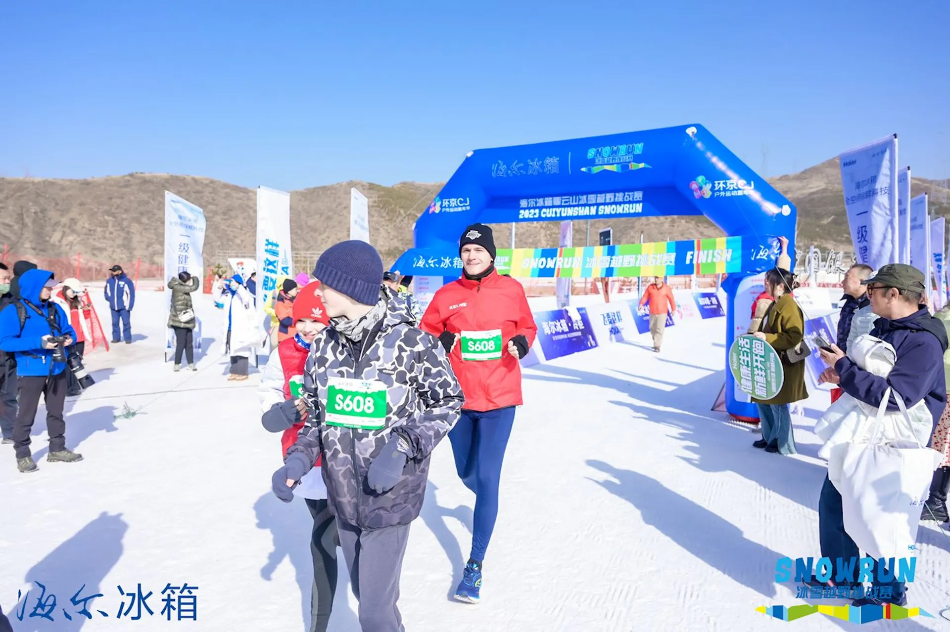 This is an image of a group of people at a snowy outdoor event, which appears to be a running race, as indicated by the "FINISH" sign at the blue archway. Participants are dressed in winter athletic gear, suitable for the cold conditions, and some of them are wearing bib numbers, a common feature in races to identify runners. In the background, there are banners with logos and what seems like sponsor names, and there's a clear blue sky, suggesting fair weather despite the snow on the ground. The atmosphere looks lively and festive with people gathered around the finish line area.