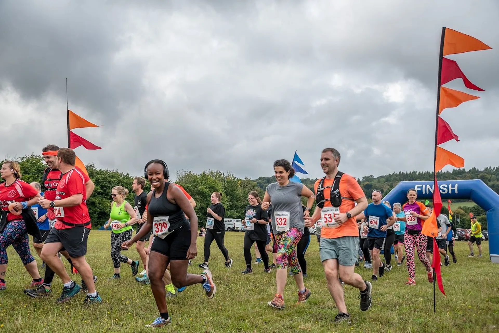 The image shows a group of people running outdoors, likely participating in a race or fun run. There are colorful flags and a "FINISH" line in the background. The participants are wearing athletic attire and have race numbers. The setting appears to be a grassy field under a cloudy sky.