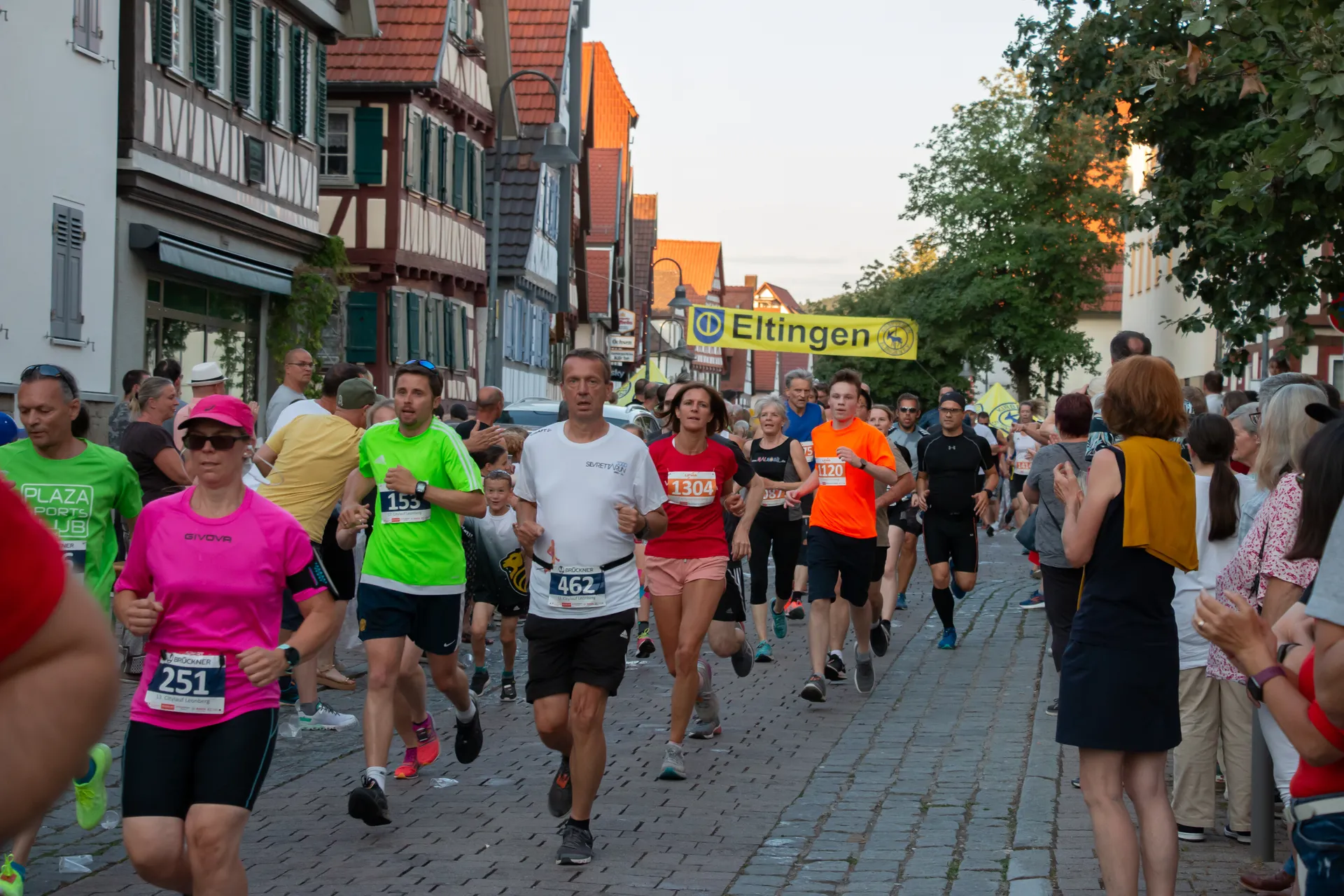The image appears to showcase a group of people participating in a road race, likely a running event such as a 5k, 10k, half-marathon, or a marathon. The participants are wearing numbers on their shirts, which are typical for such events to identify the runners. They are running on a street lined with spectators who are watching and possibly cheering them on. The buildings along the street have a traditional European architectural style, indicating that this event might be taking place in a town in Europe. The weather looks fair, and it seems to be either in the late afternoon or early evening, considering the natural lighting.